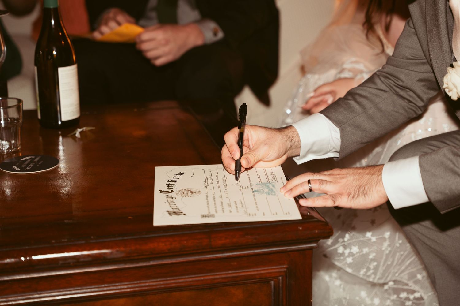 Close-up of hands signing wedding documents on dark wooden table.