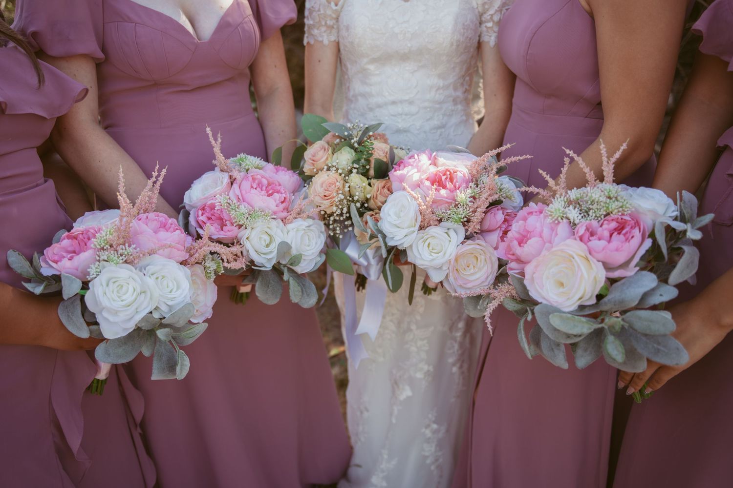 Row of bridesmaids holding pink and white floral bouquets against natural backdrop.