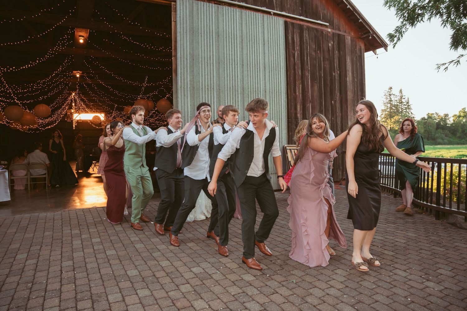 Group performs coordinated dance moves at an outdoor wedding reception with string lights overhead.
