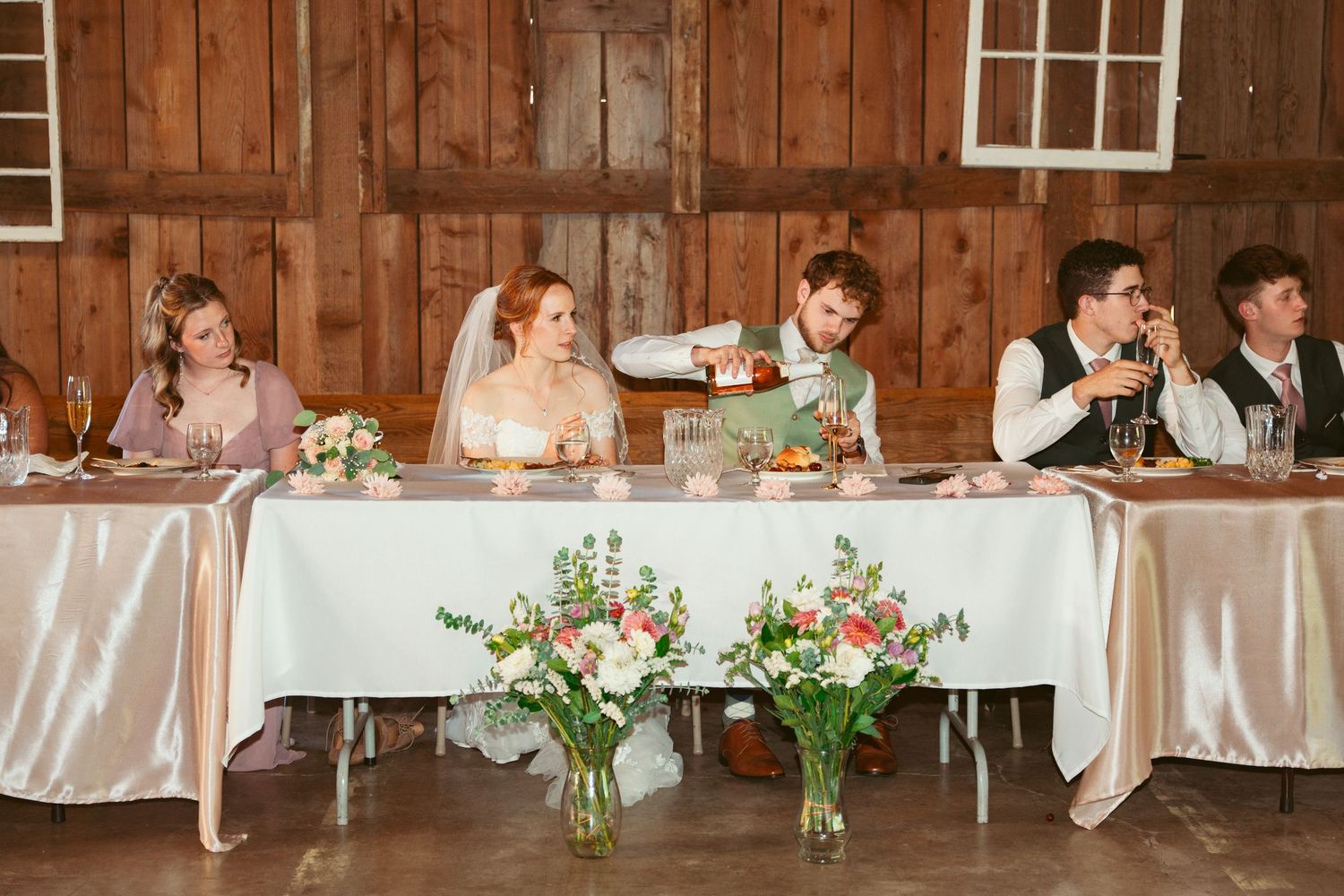 Wedding party seated at a long head table decorated with floral arrangements in a rustic barn setting.
