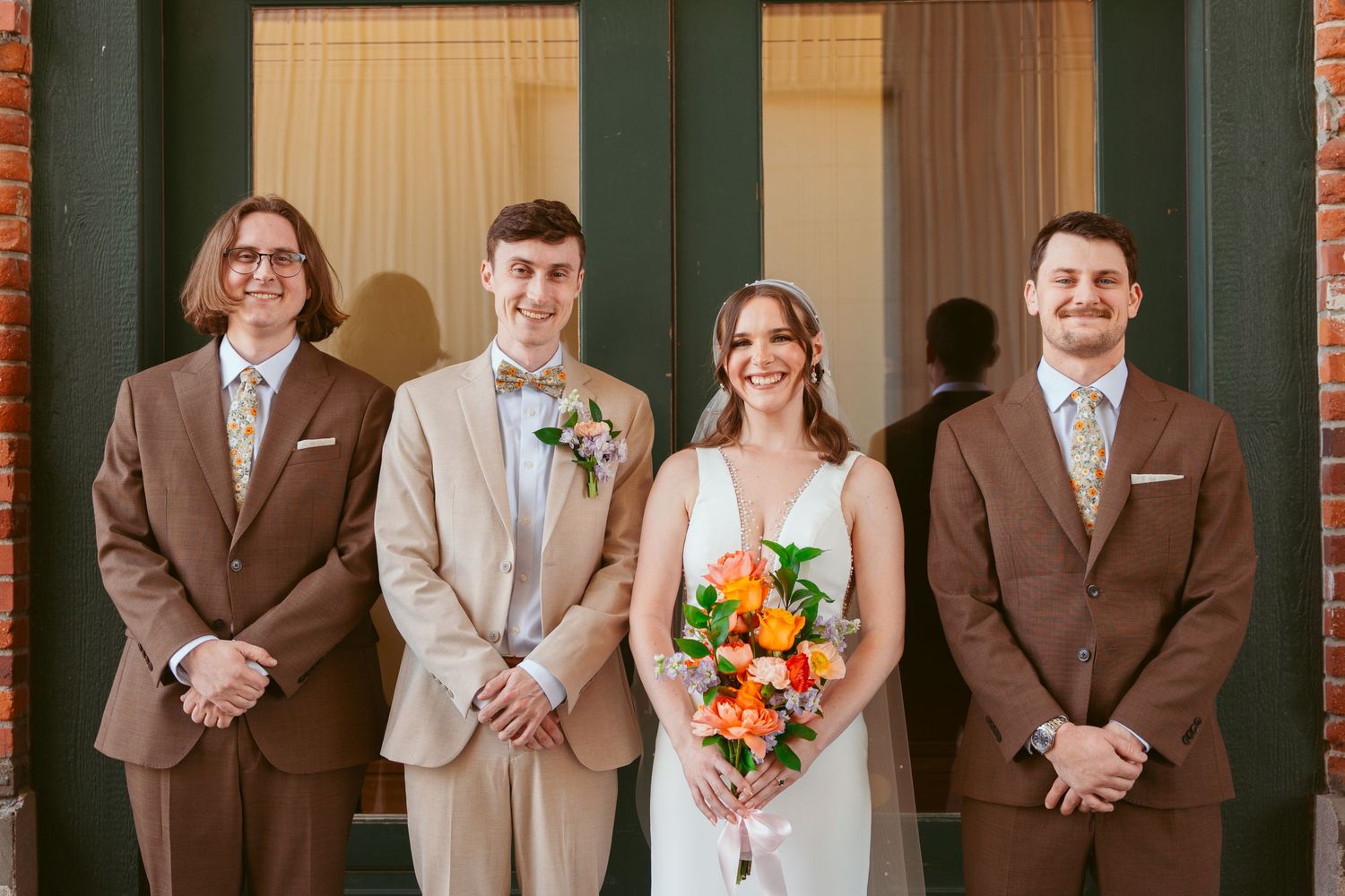 Wedding party group shot features brown suits, light dresses, and bright floral bouquets.