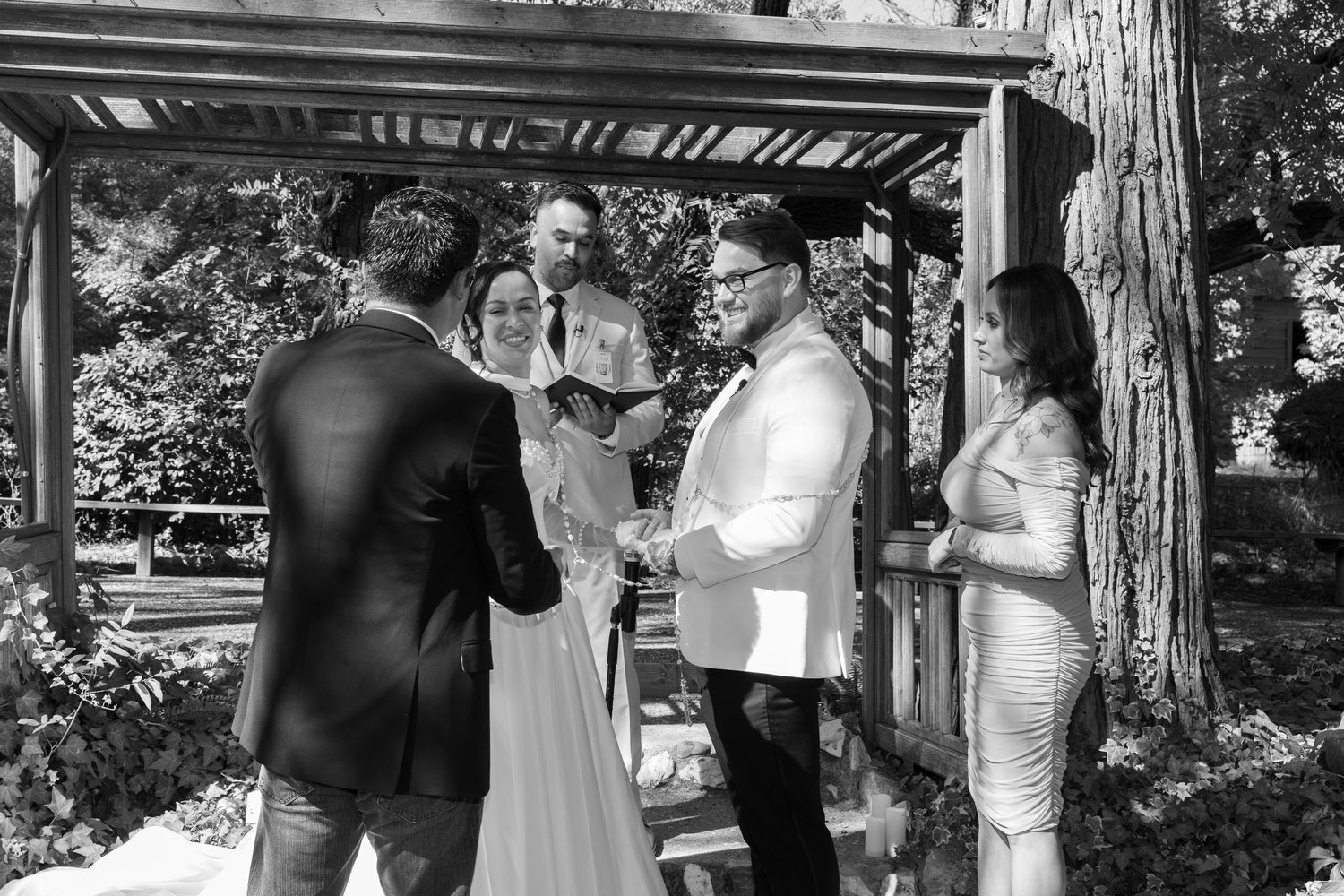 Black and white photo of wedding ceremony under rustic wooden structure with natural lighting.