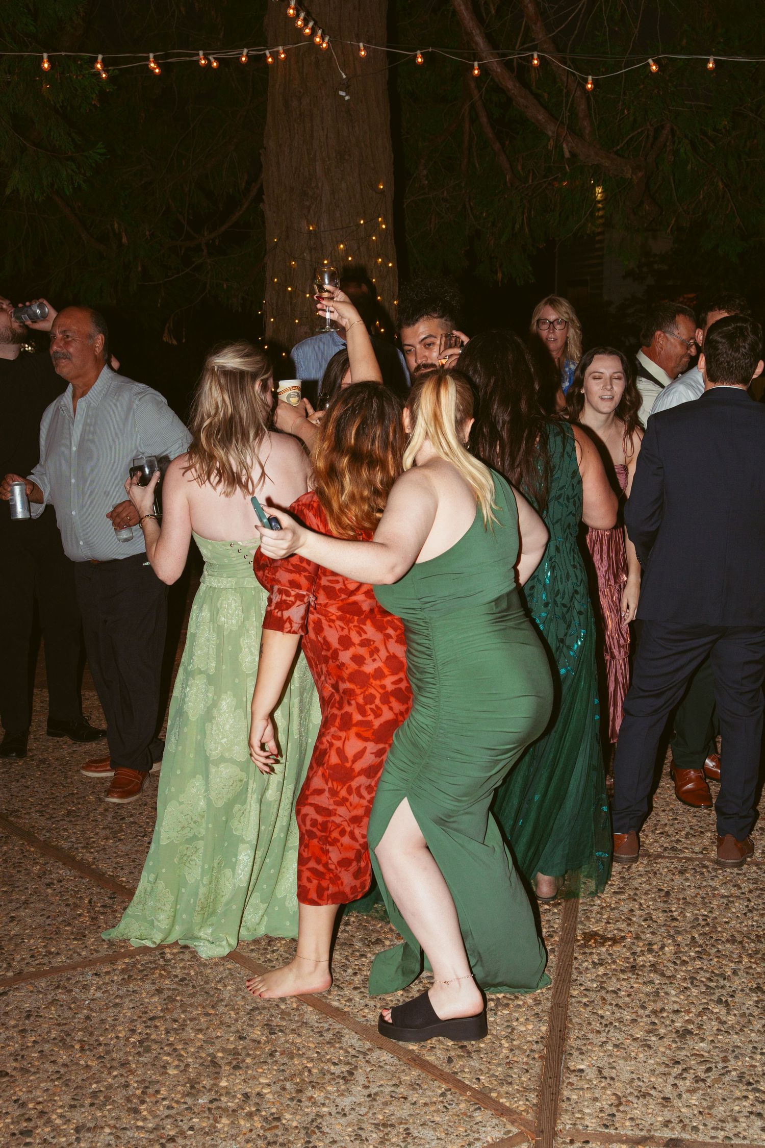 Wedding guests in colorful dresses dance together on a gravel dance floor under twinkling lights.