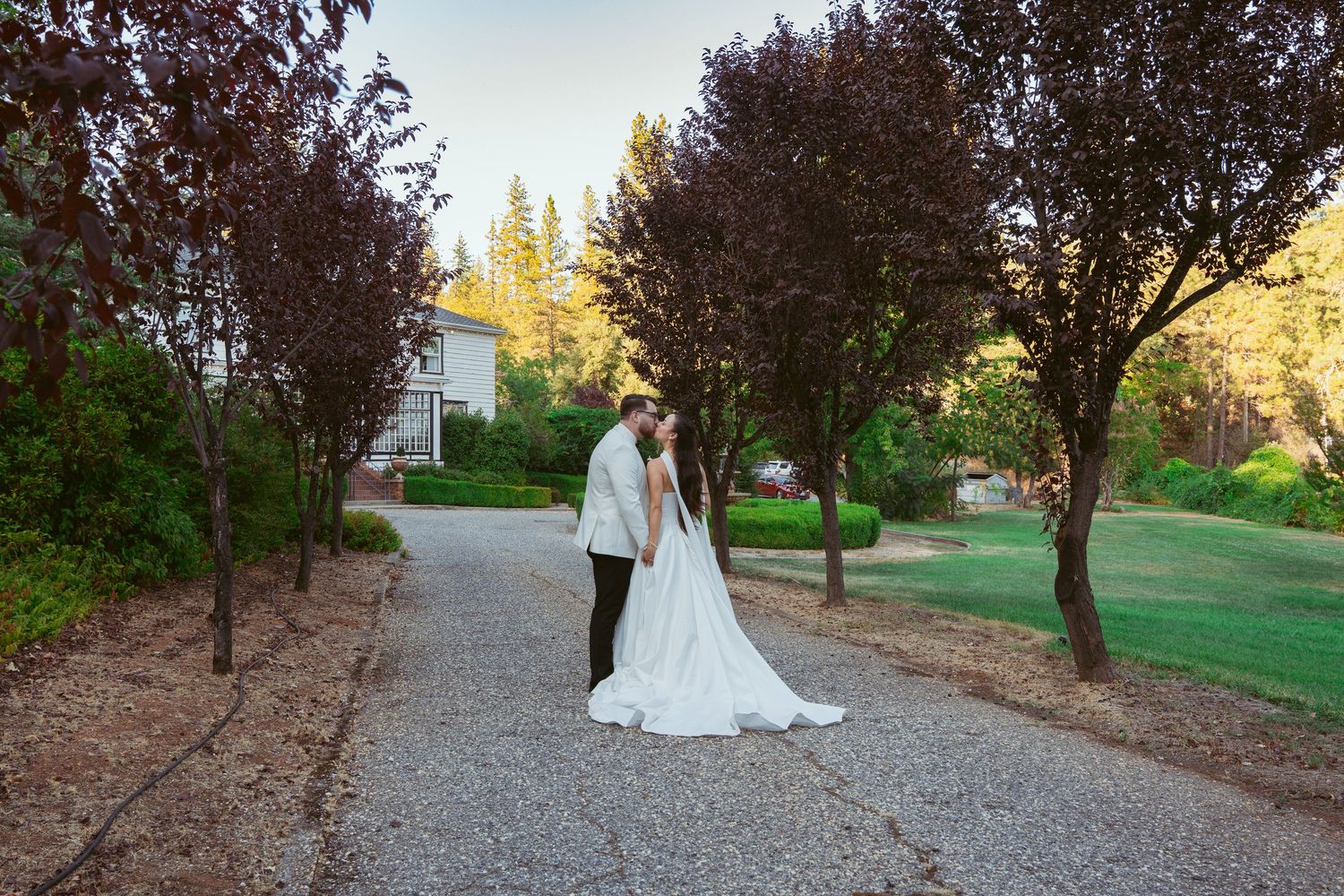 A couple shares a romantic moment on a gravel path surrounded by trees at sunset during their wedding celebration.