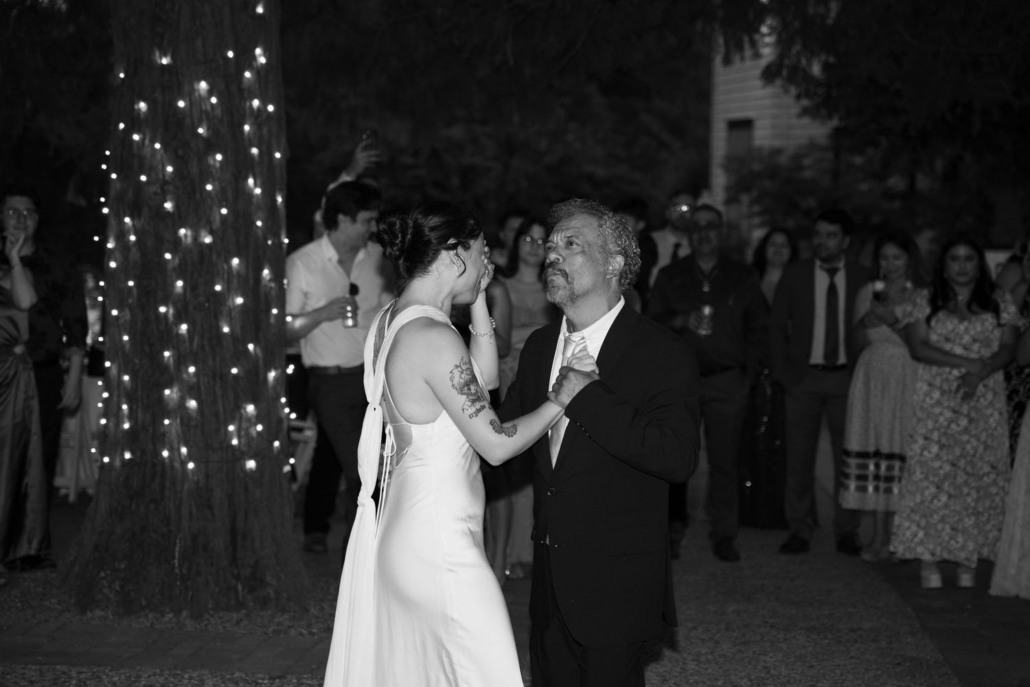 Wedding guests dance under glowing string lights at an evening outdoor reception.