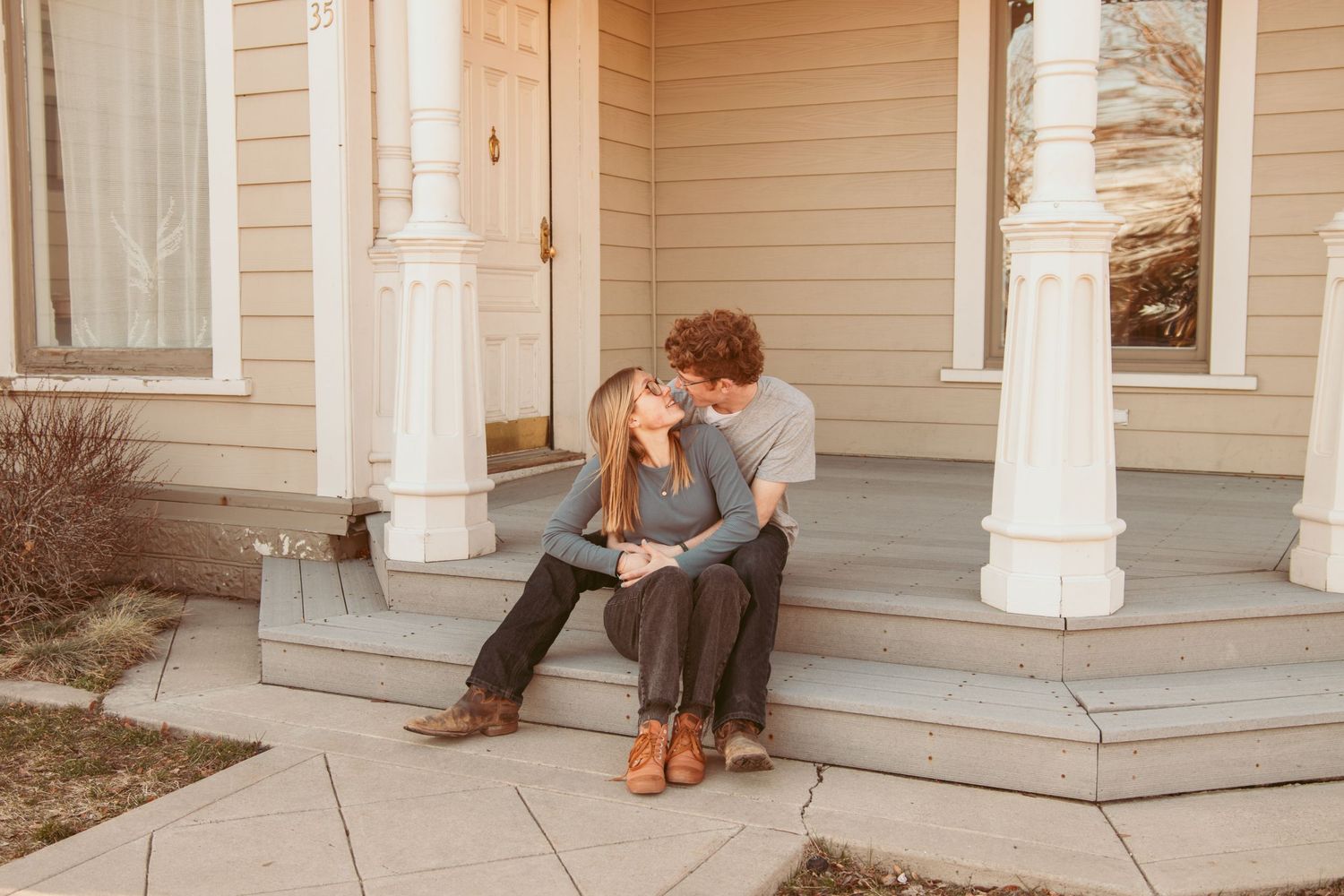 A romantic couple shares tender moments on a sunny porch with white columns and beige siding.