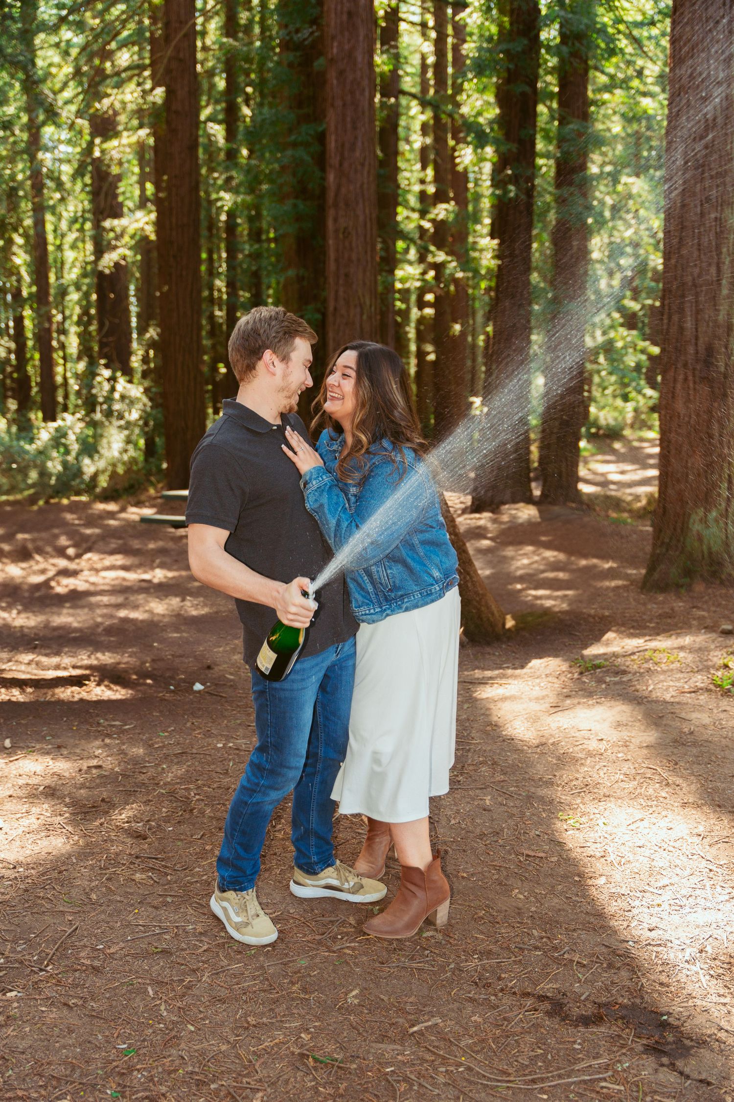 Champagne sprays as a couple celebrates joyfully among tall redwood trees.