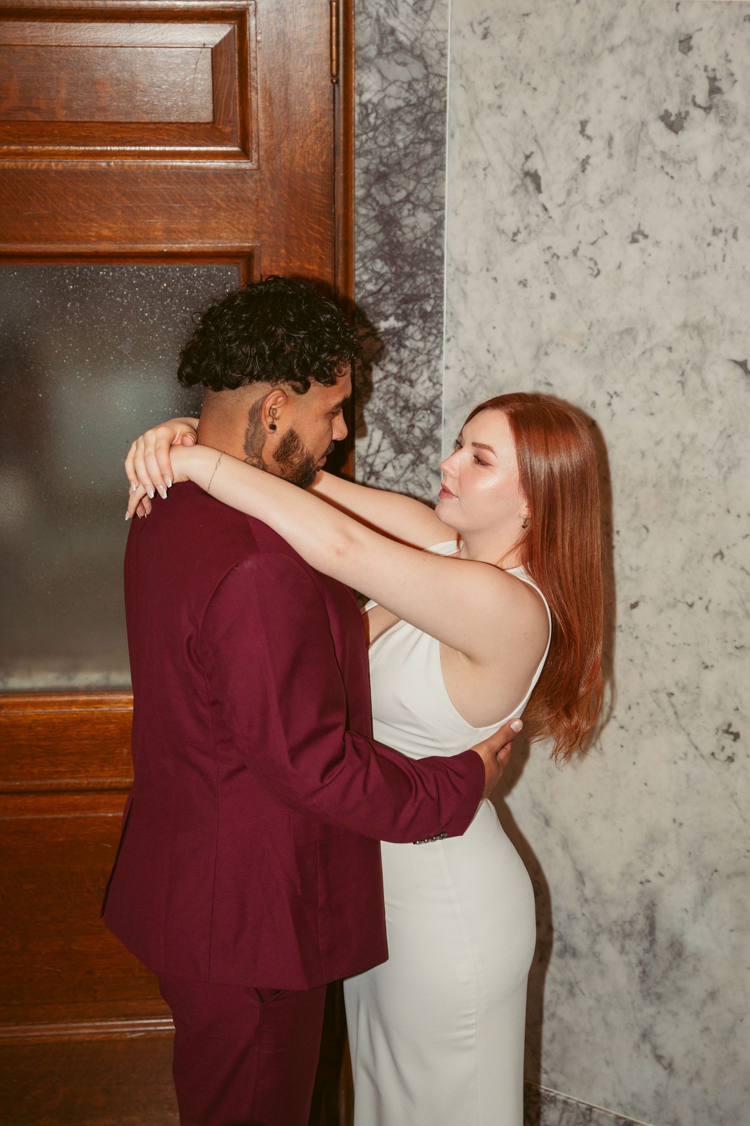 An intimate embrace between a couple in burgundy and white against a wooden door and marble wall.