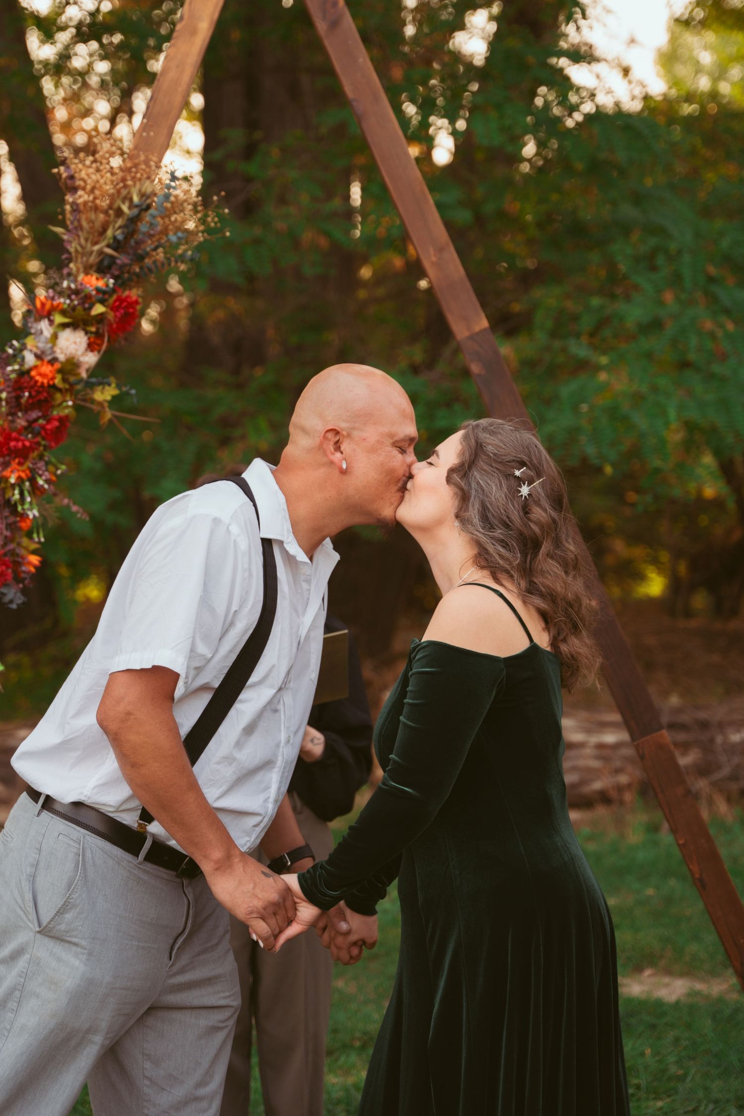 A romantic first kiss between newlyweds under a wooden arch decorated with red flowers.