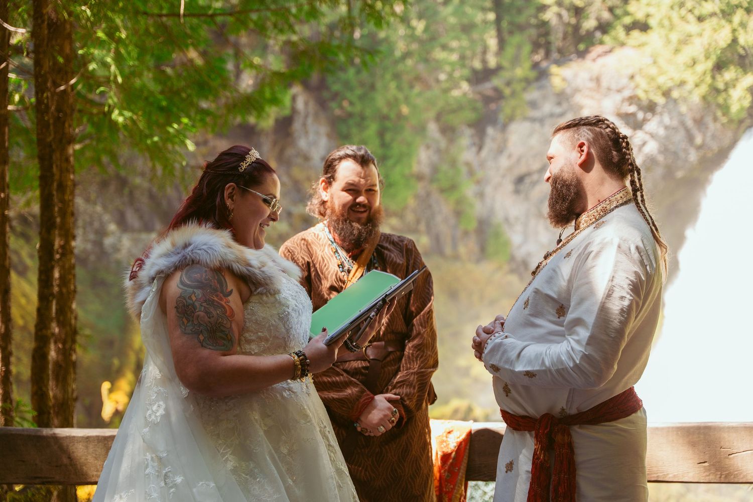 A wedding ceremony takes place among tall trees in a sunlit forest setting.
