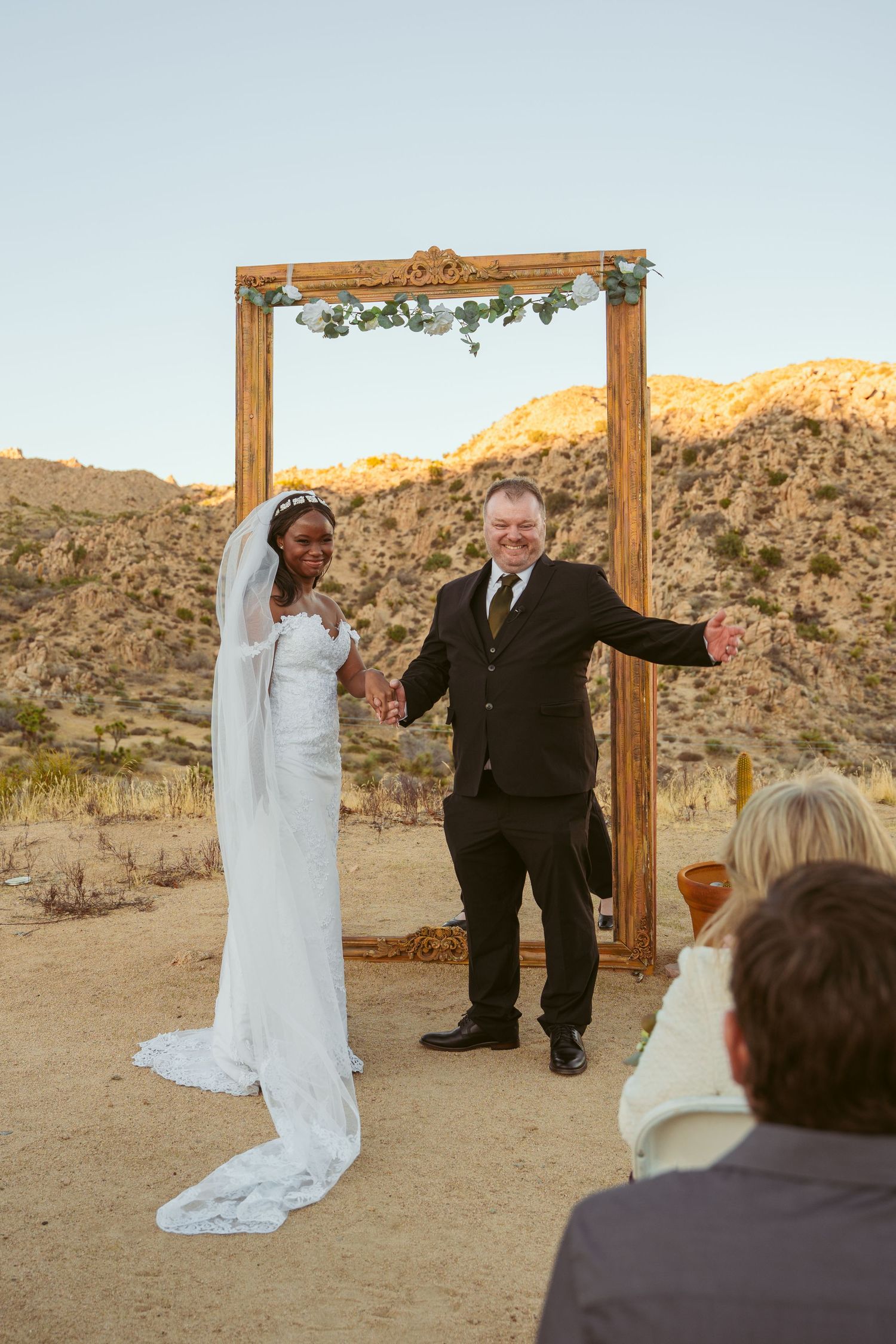 A desert wedding ceremony setup with a wooden arch frame and chairs against a mountainous backdrop at sunset.