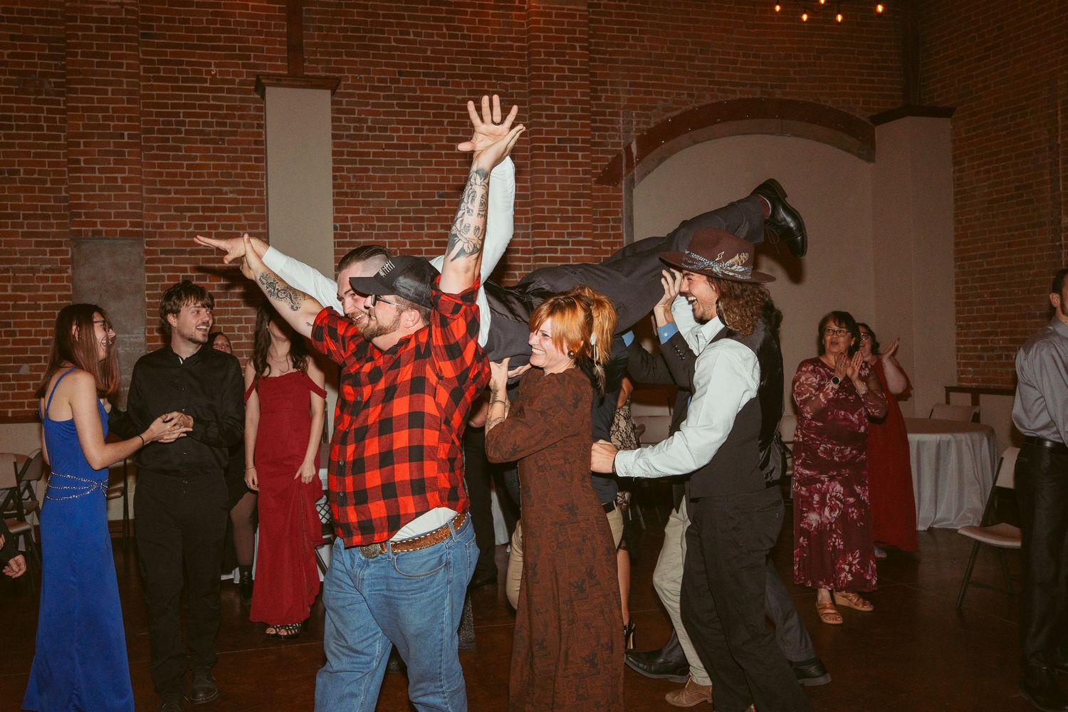Festive crowd dancing beneath string lights in a brick-walled venue with formal attire.