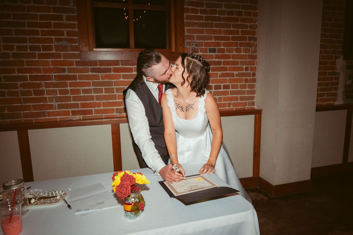 Newlyweds share a kiss while signing their marriage license at a reception table with red brick wall backdrop.