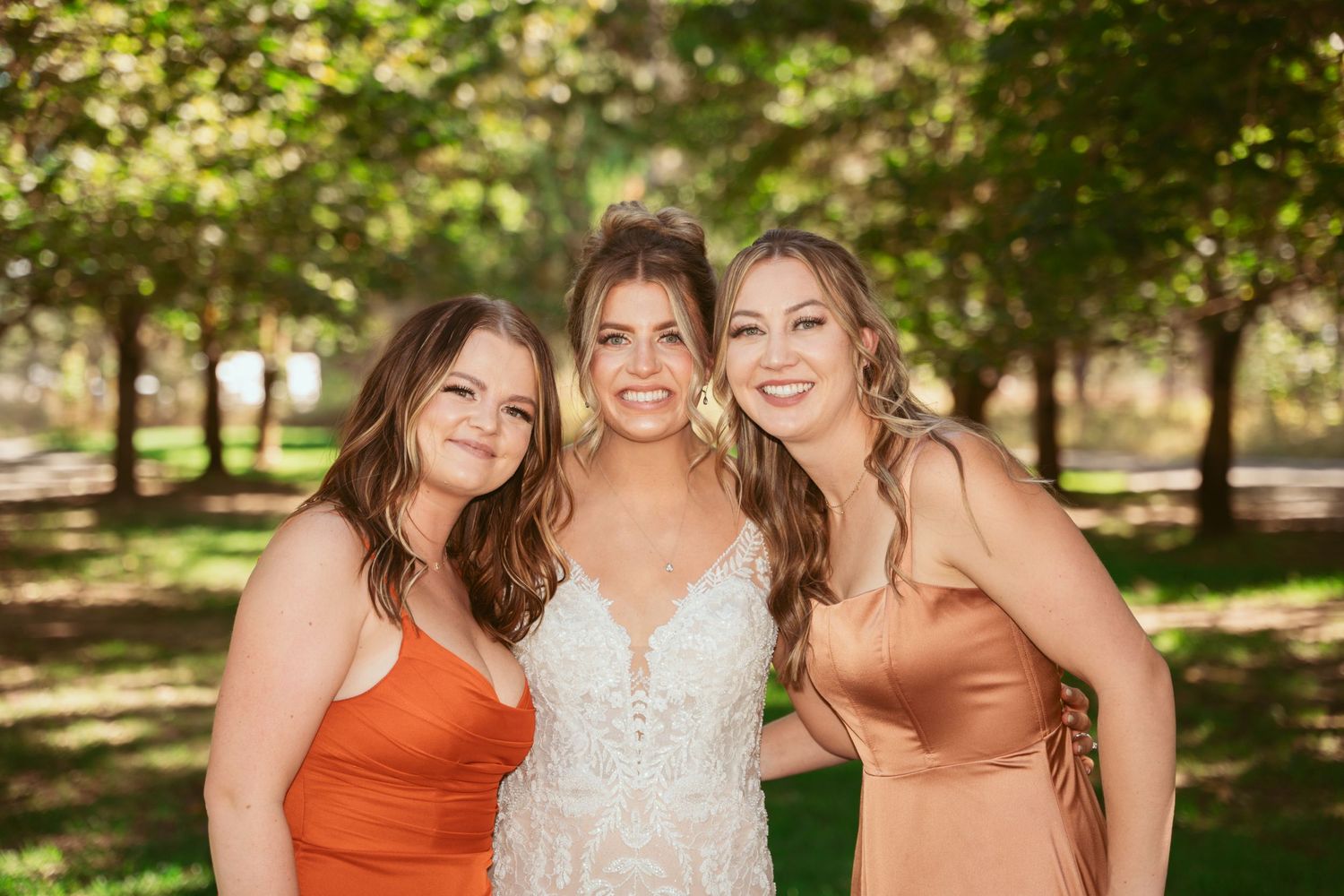 Three women in formal attire pose together outdoors on a sunny day with trees in the background.