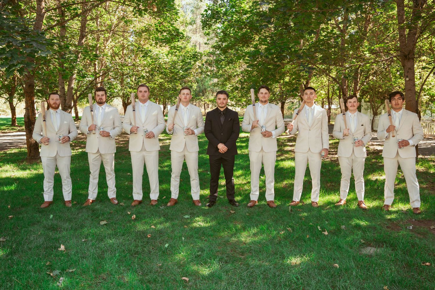 Groomsmen in crisp white suits pose with person in black at center in sunny garden setting.
