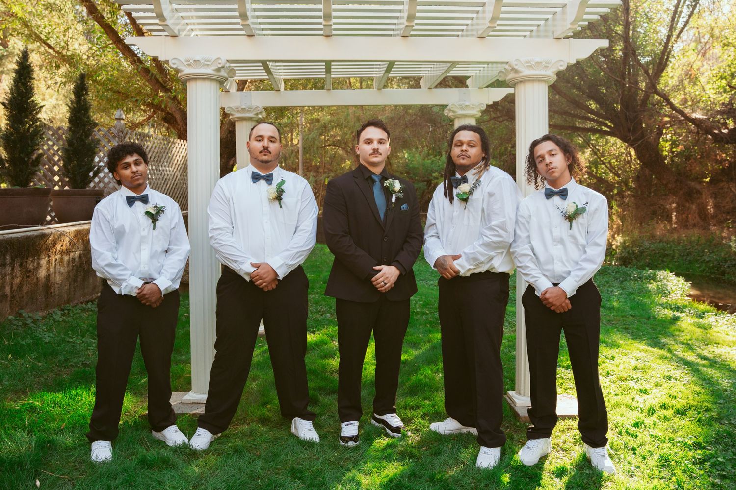 Groomsmen in black suits pose playfully on the ground under a white pergola.