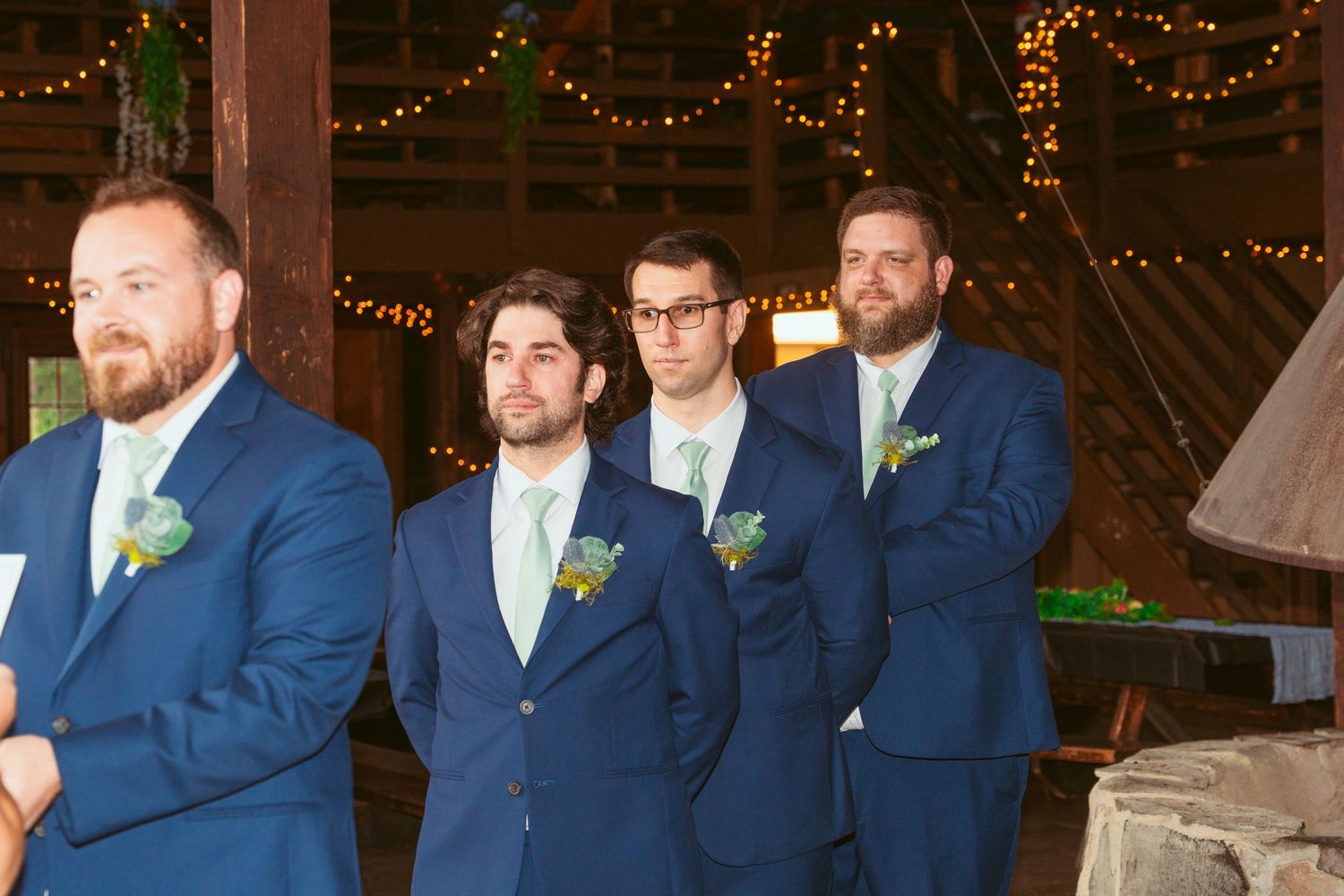 Groomsmen in navy suits line up inside barn with string lights overhead.