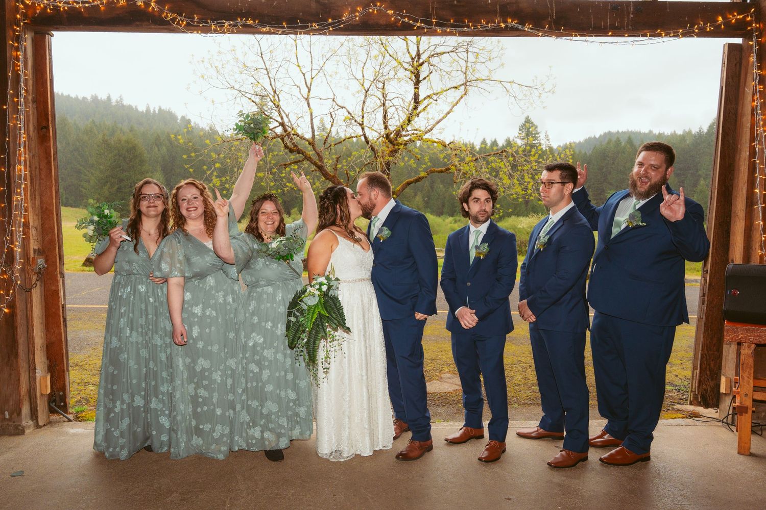 Wedding party laughs together while posing under rustic wooden arch with mountain backdrop.
