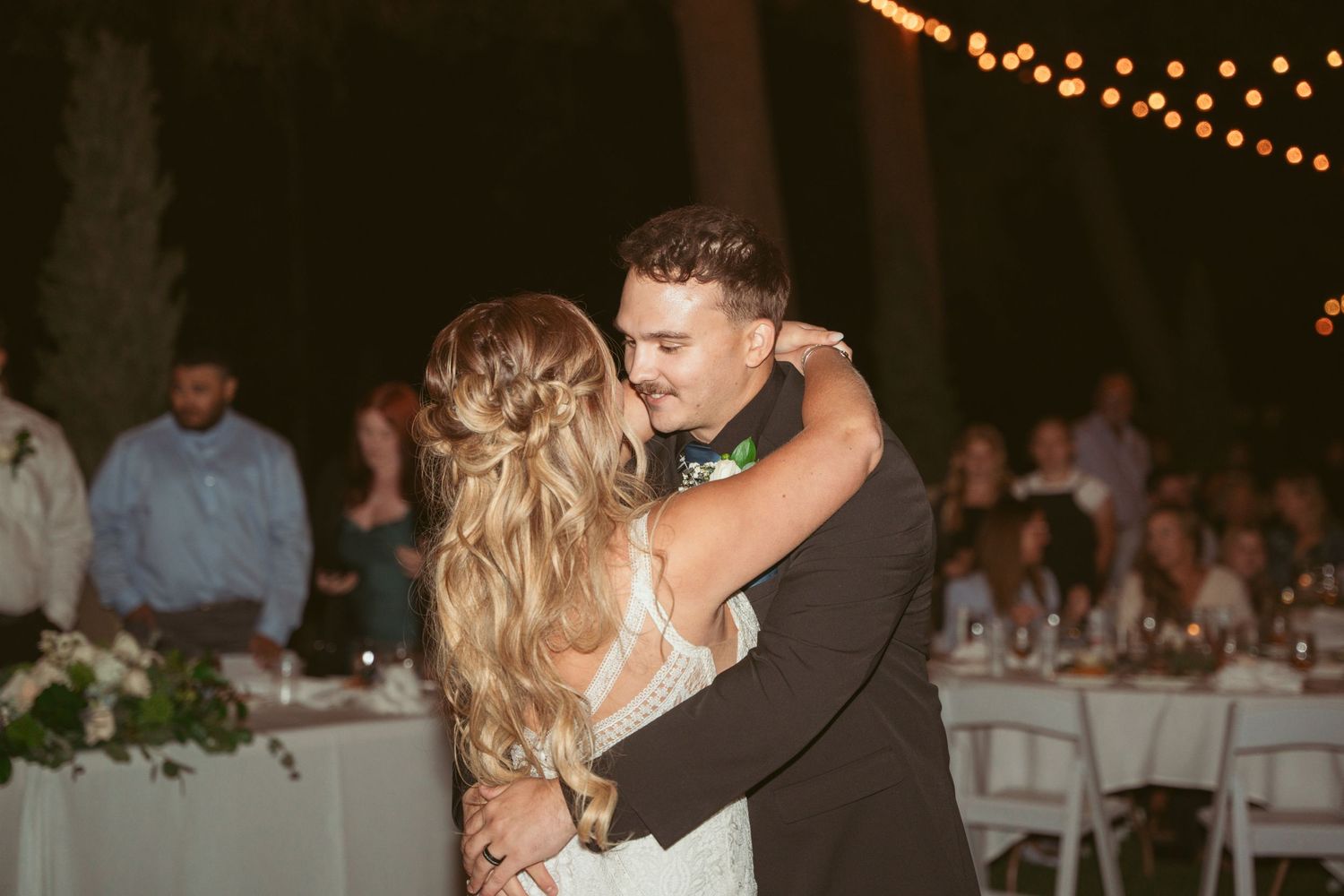 Couples slow dance beneath warm string lights at a romantic outdoor wedding reception.