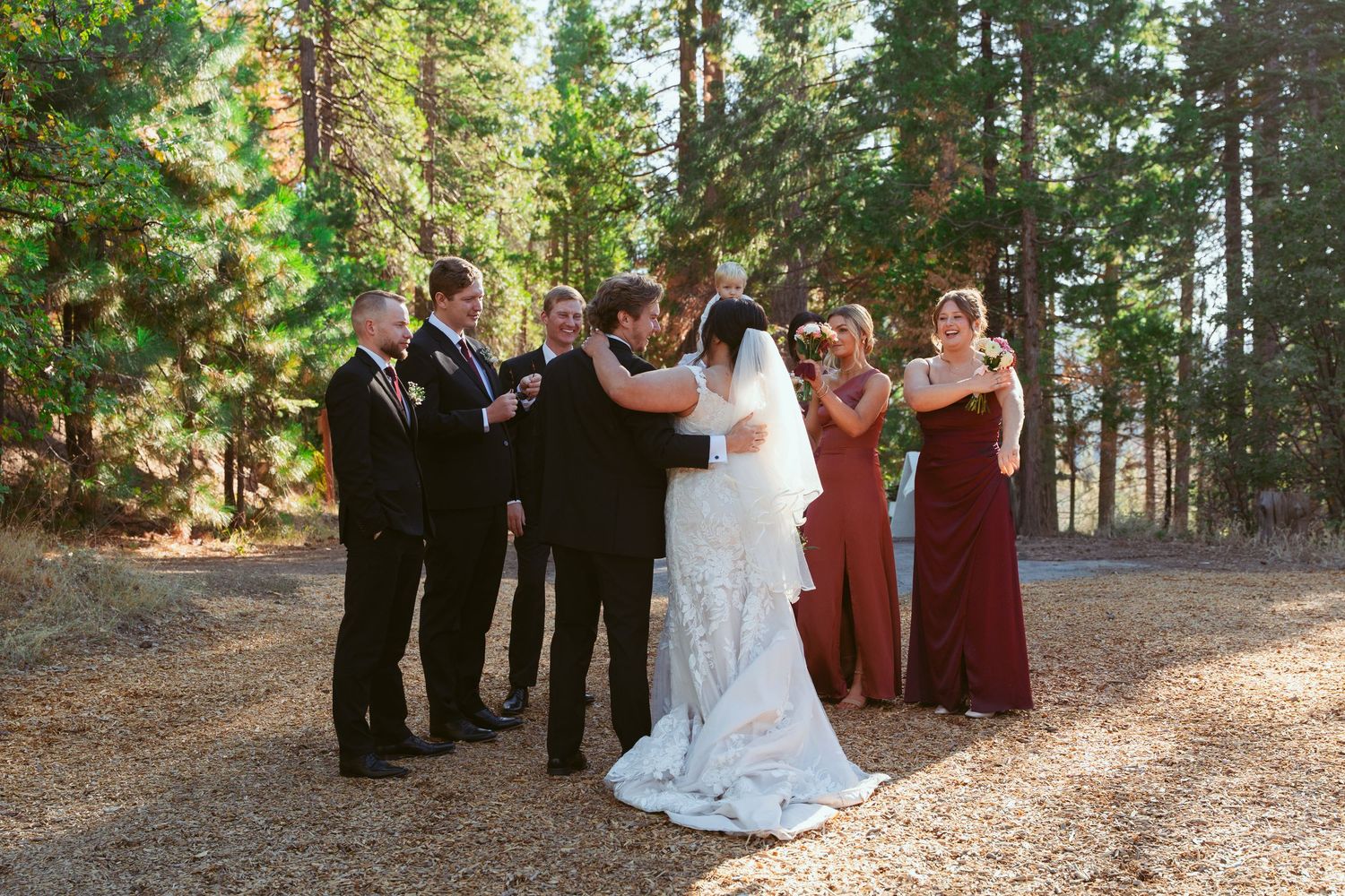 An outdoor forest wedding ceremony with attendees in formal attire and burgundy bridesmaid dresses.