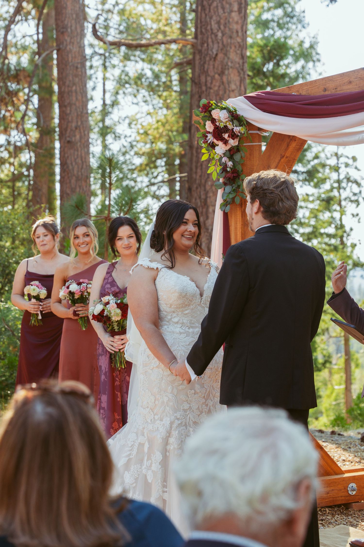 A wedding ceremony takes place under tall pine trees with a wooden arch and burgundy drapery.