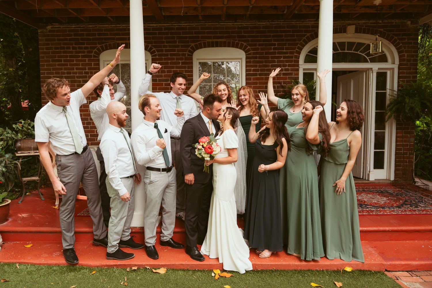 Wedding party celebrating on a red-carpeted porch of a brick building with mixed formal attire.