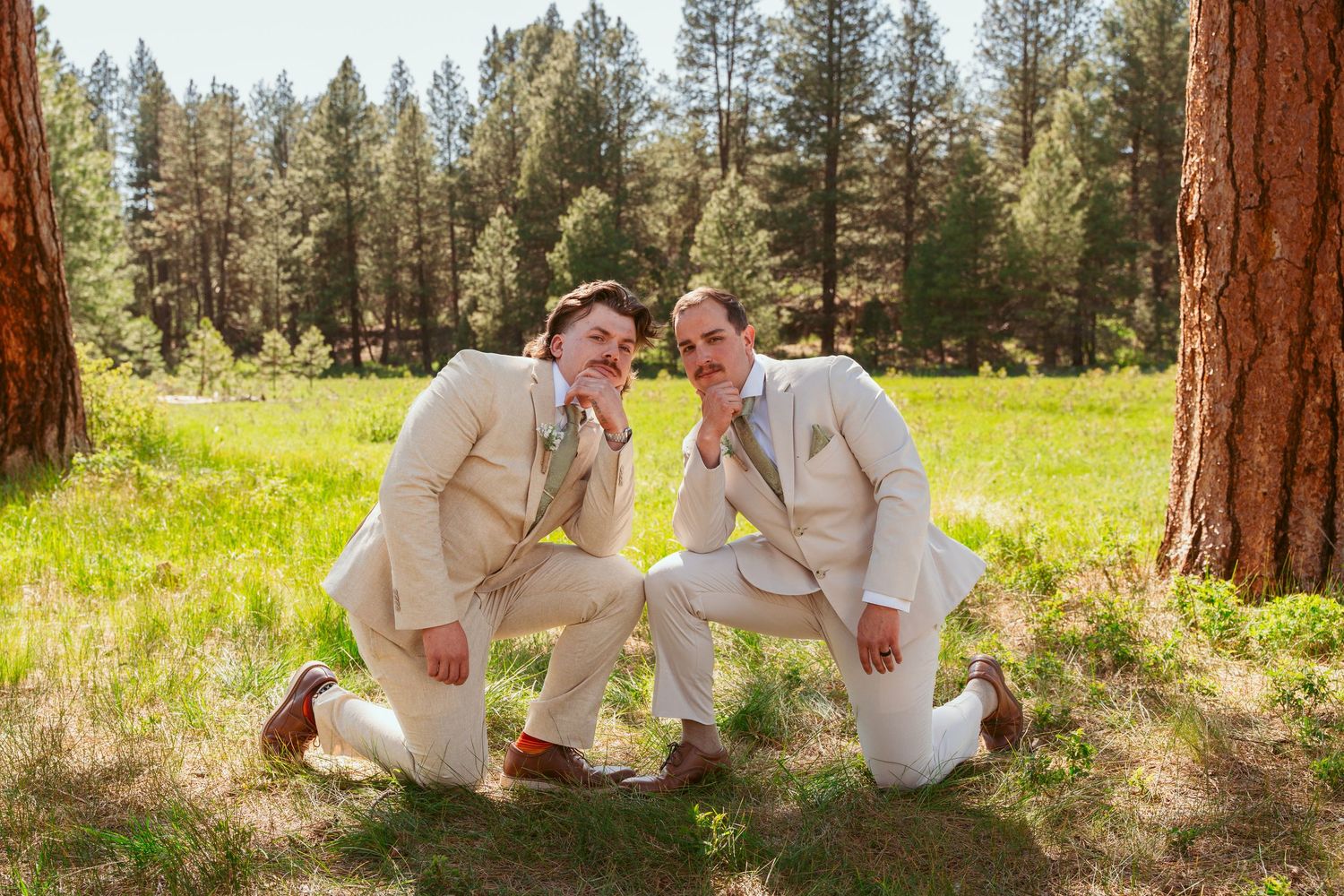 Wedding party members in cream suits relax casually against pine trees in a meadow.