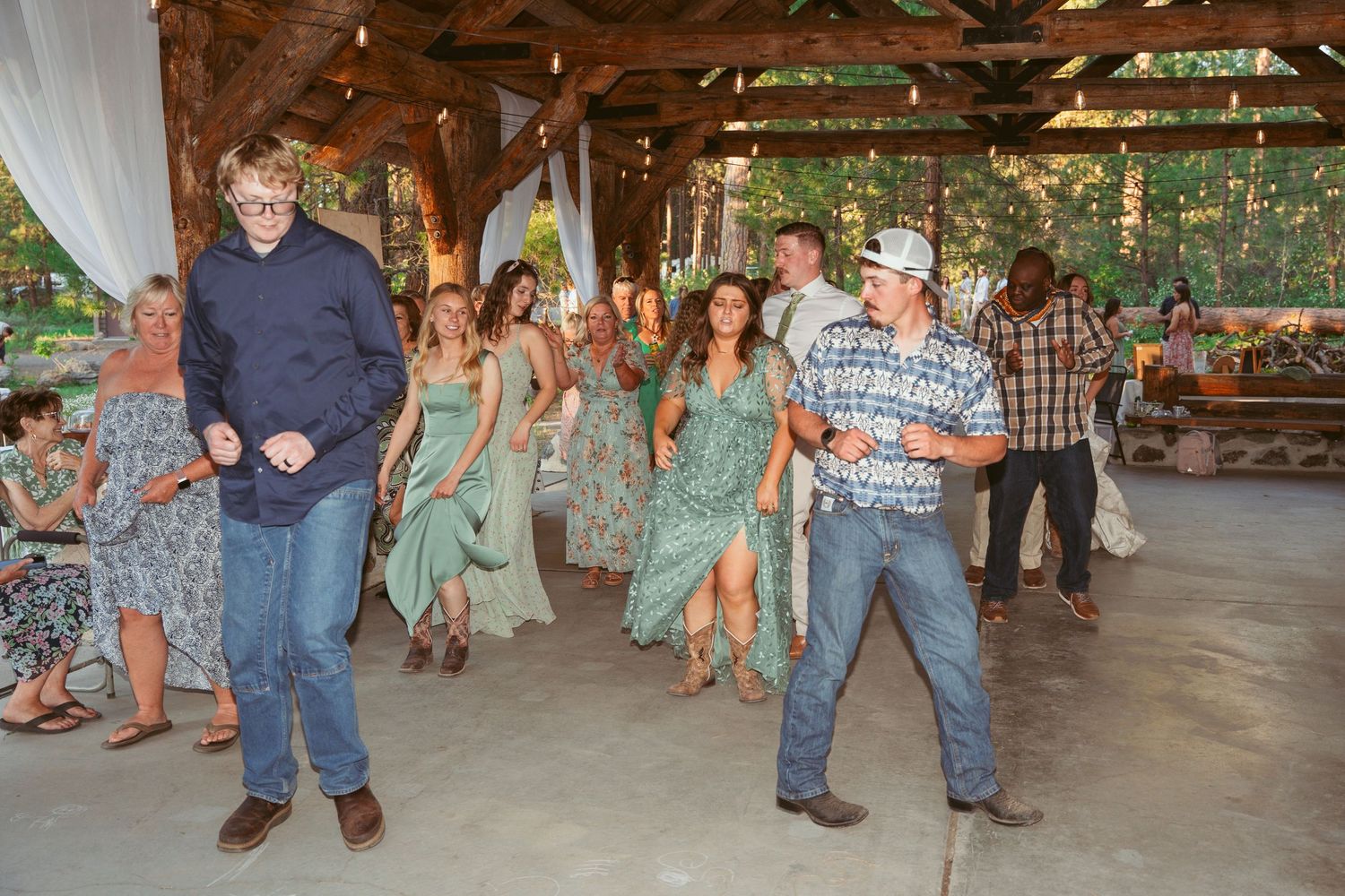 Wedding guests performing group dances on a wooden dance floor under string lights.