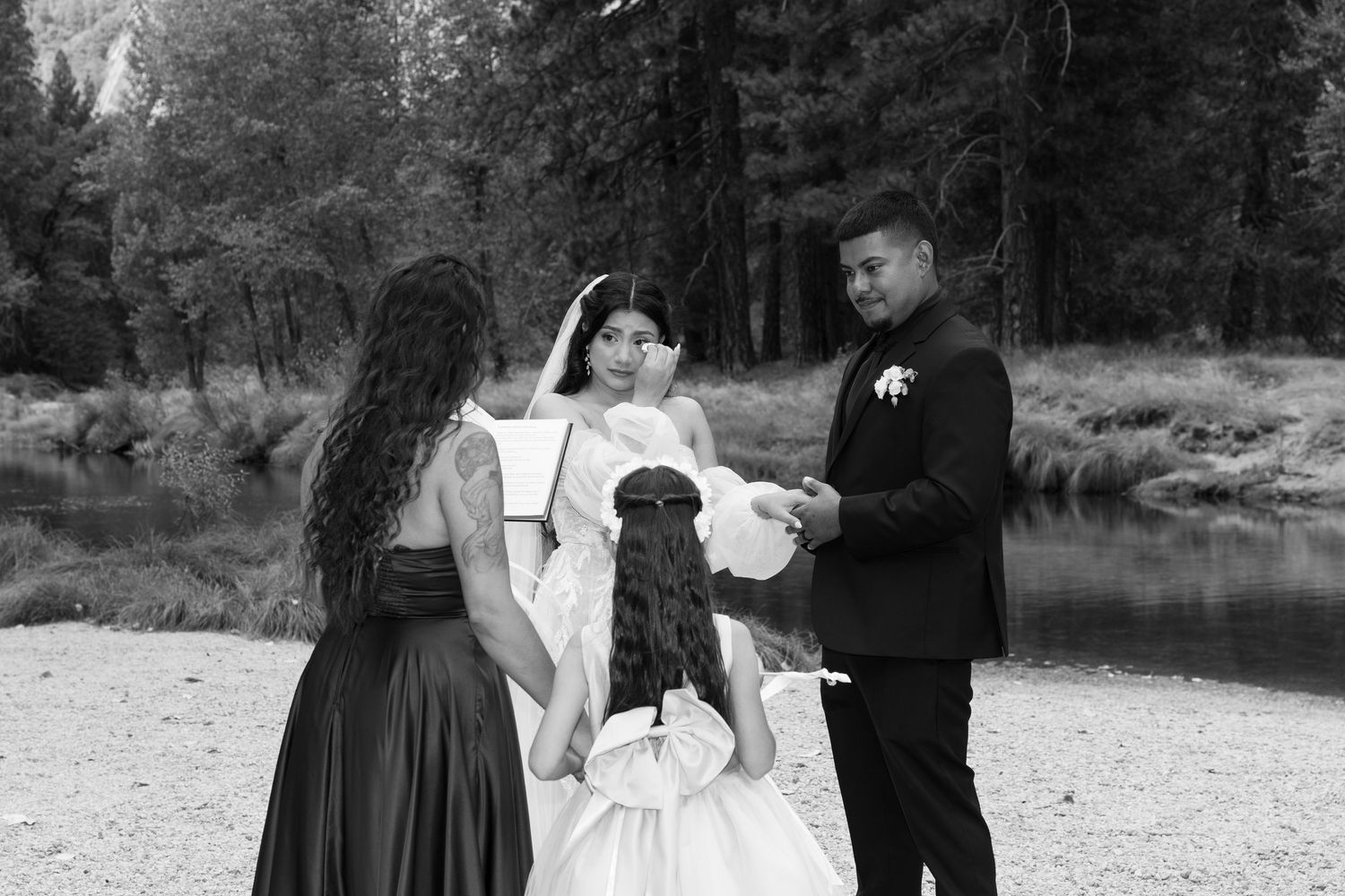 Black and white photo of wedding party gathered on rocky riverbank in forest setting.