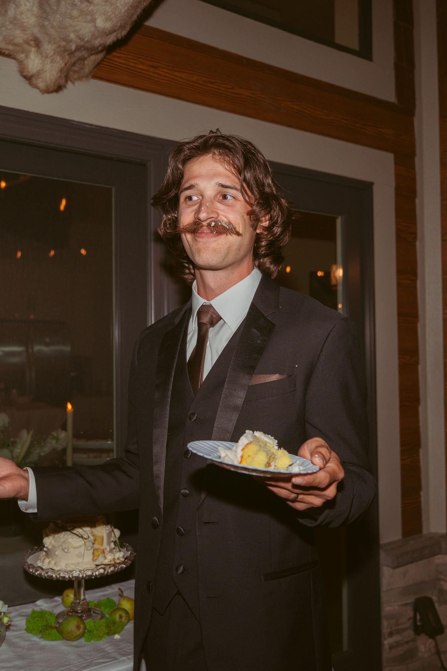 A groom with a mustache smiles while holding wedding cake at the reception.