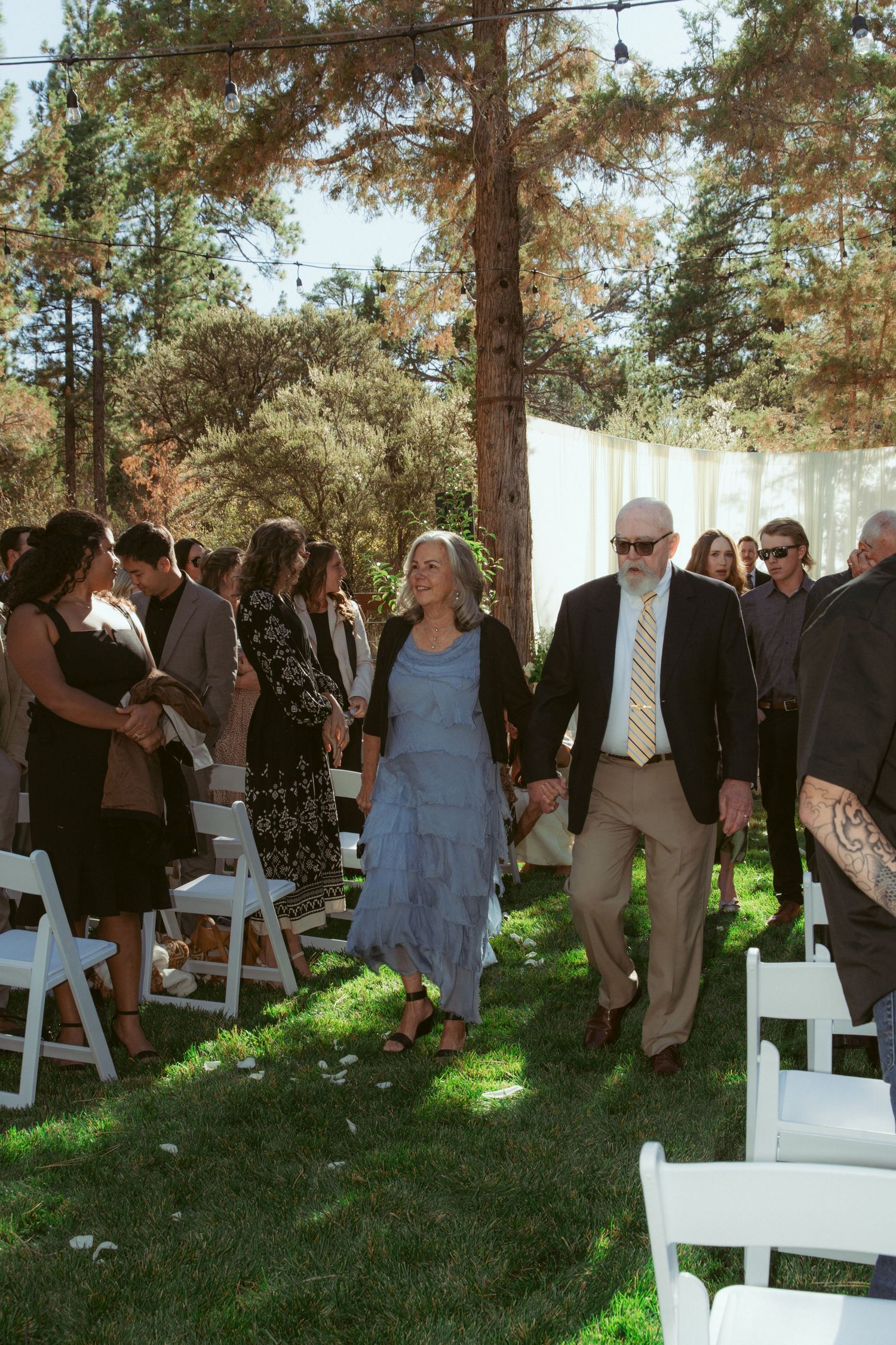 Wedding guests and bridal party socialize before ceremony on grassy area with white chairs and pine trees.