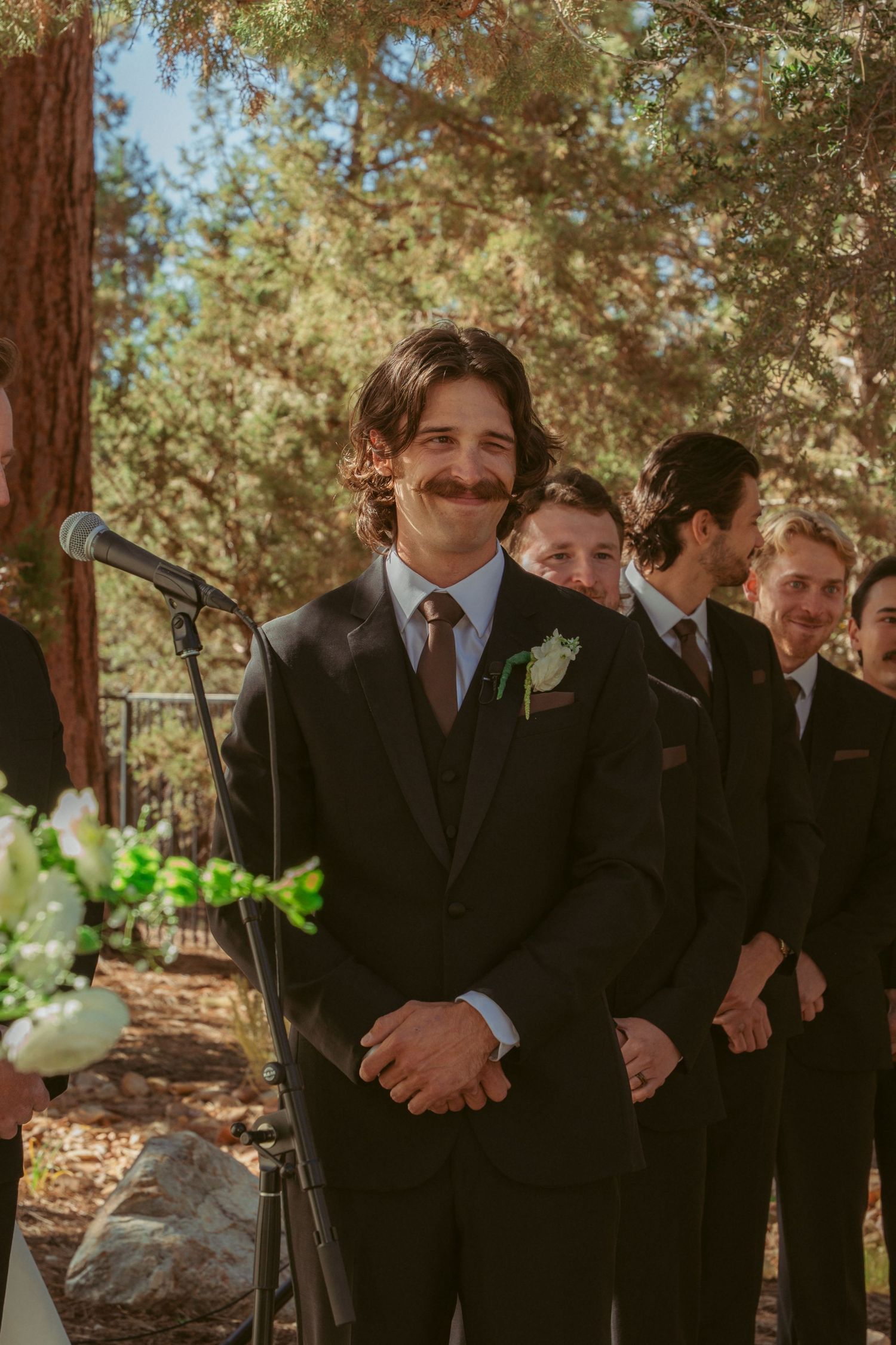 Groomsmen in dark suits pose together under pine trees at outdoor wedding venue.