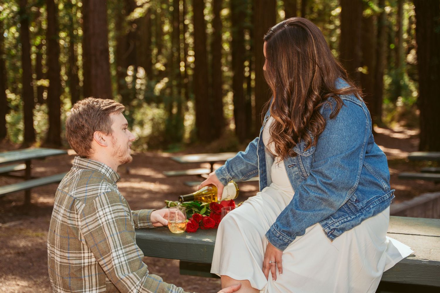 A romantic couple shares a tender moment on a wooden bench in a sunlit forest setting.