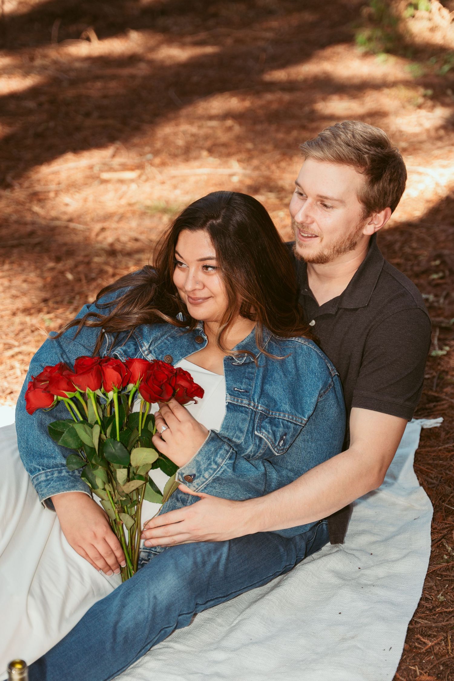 Romantic forest picnic scene with couple holding red roses on white blanket.