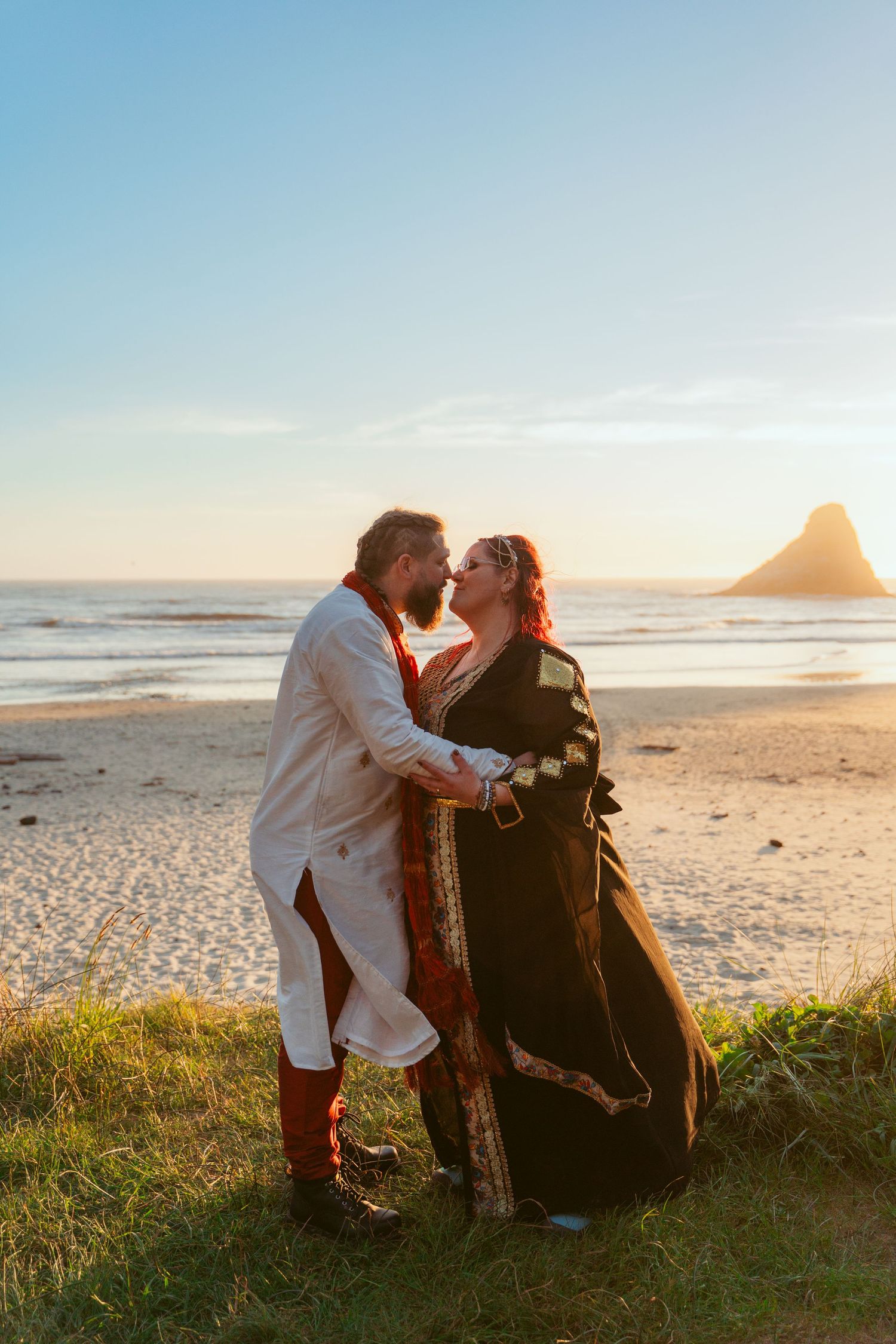 A tender moment between newlyweds in medieval-inspired attire against a coastal sunset backdrop.