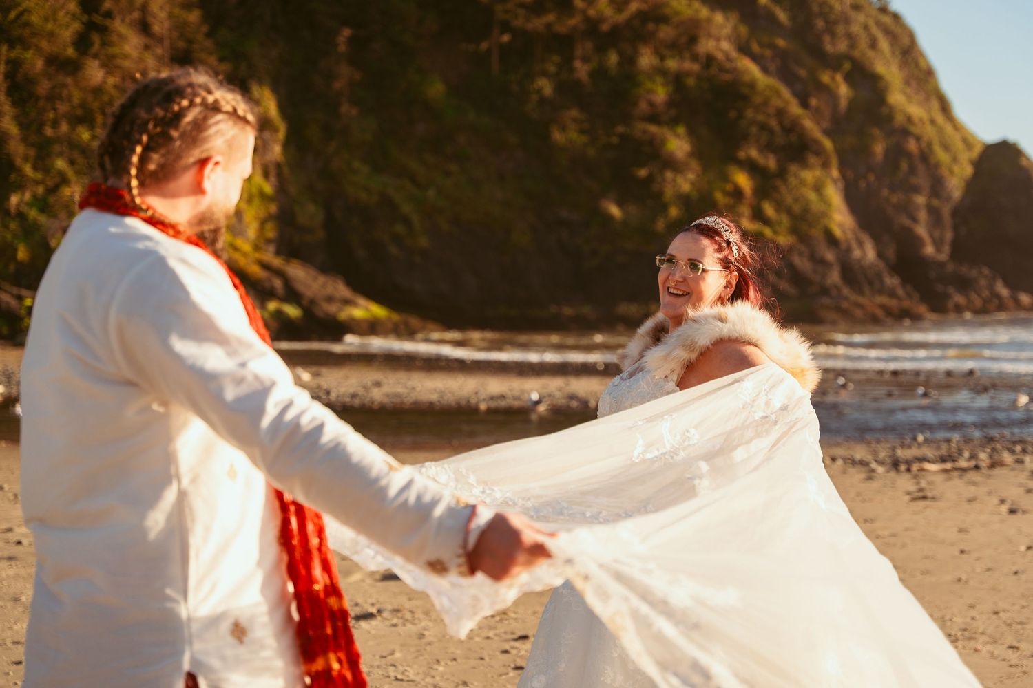 Two people in contrasting red and white outfits twirl playfully on a sandy beach near a coastal cliff.