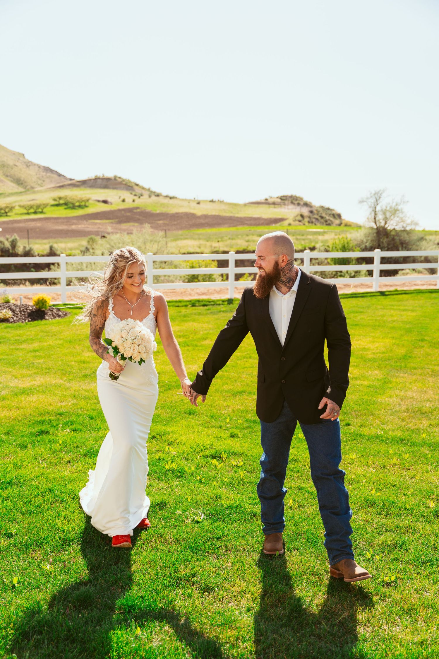 Newlyweds playfully chase each other across a grassy field with white fencing and hills in the background.
