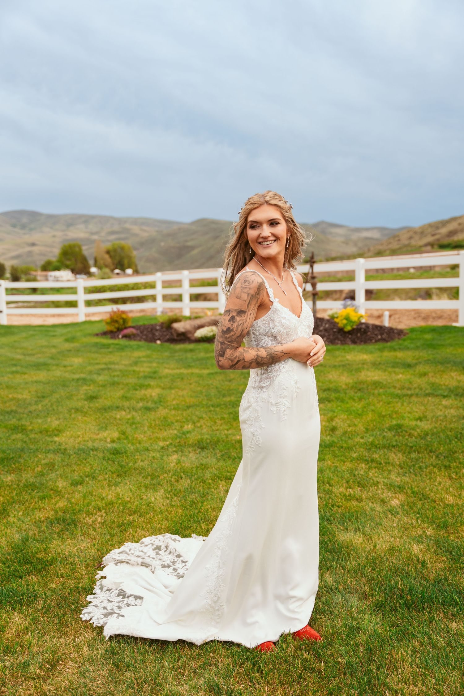 Bride in flowing white dress twirls on green lawn with white fence and mountains in background.