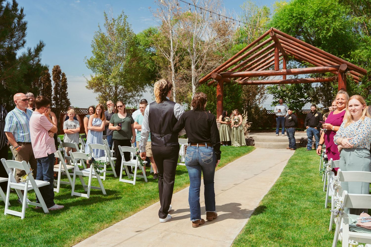Wedding ceremony setup features rows of white chairs leading to wooden pergola on manicured lawn.