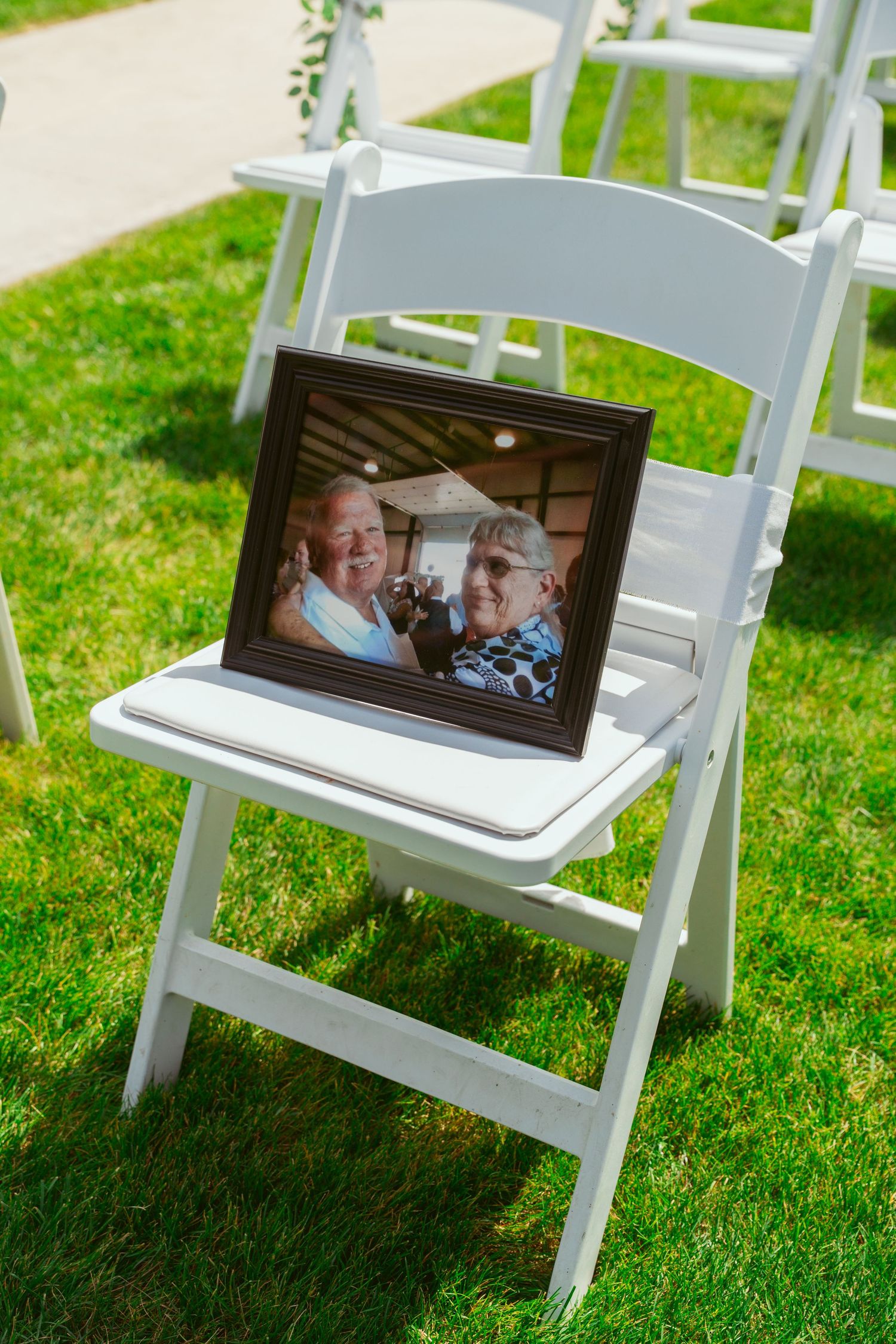 White folding chairs arranged on grass with framed photos placed on seats during an outdoor event.