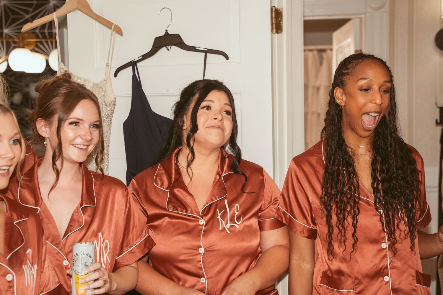 Wedding party members in matching silk pajamas share laughs and drinks during pre-ceremony preparations.
