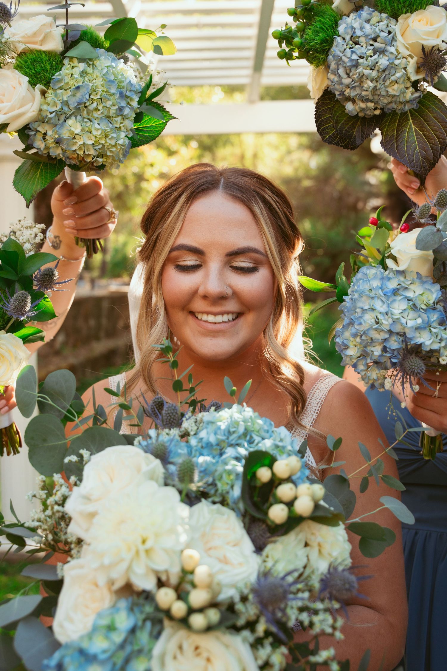 Bride surrounded by blue hydrangea and white rose bouquets smiles with eyes closed in garden setting.
