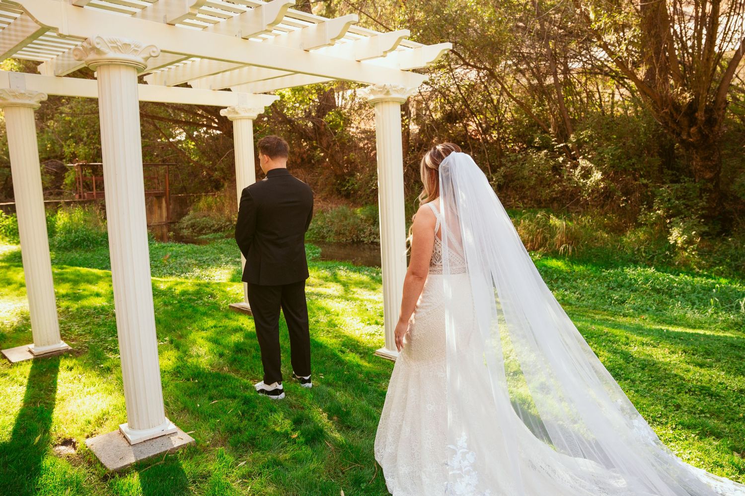 Wedding ceremony taking place under a white pergola with sunlit grass in the background.