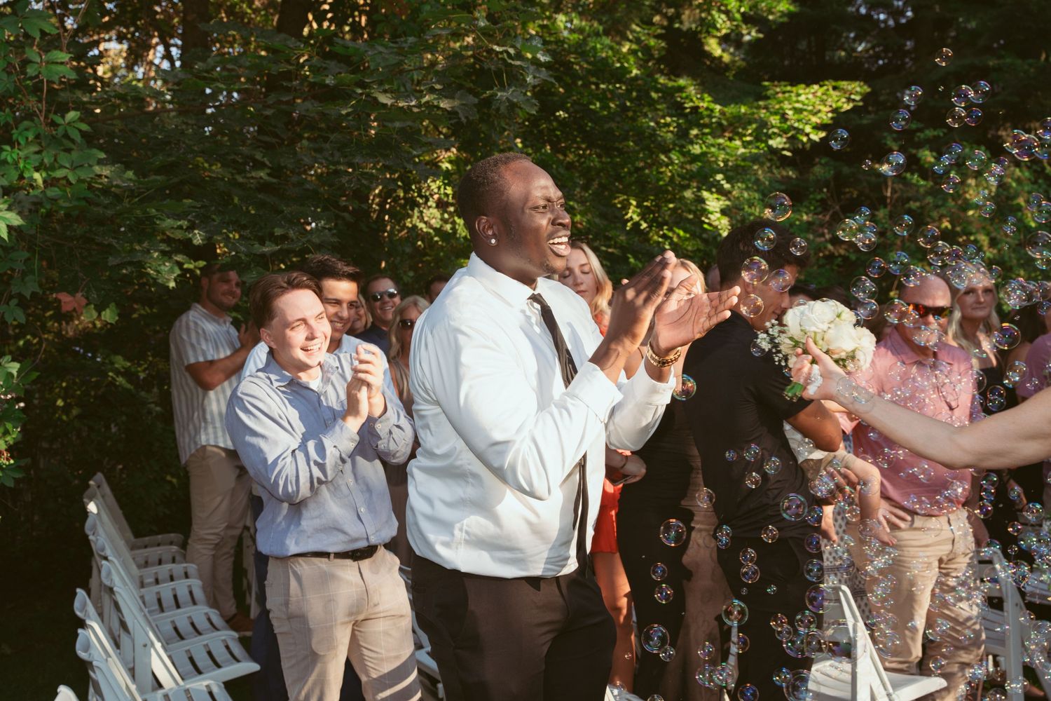 Joyful wedding celebration with guests dancing outdoors under green tree canopy.