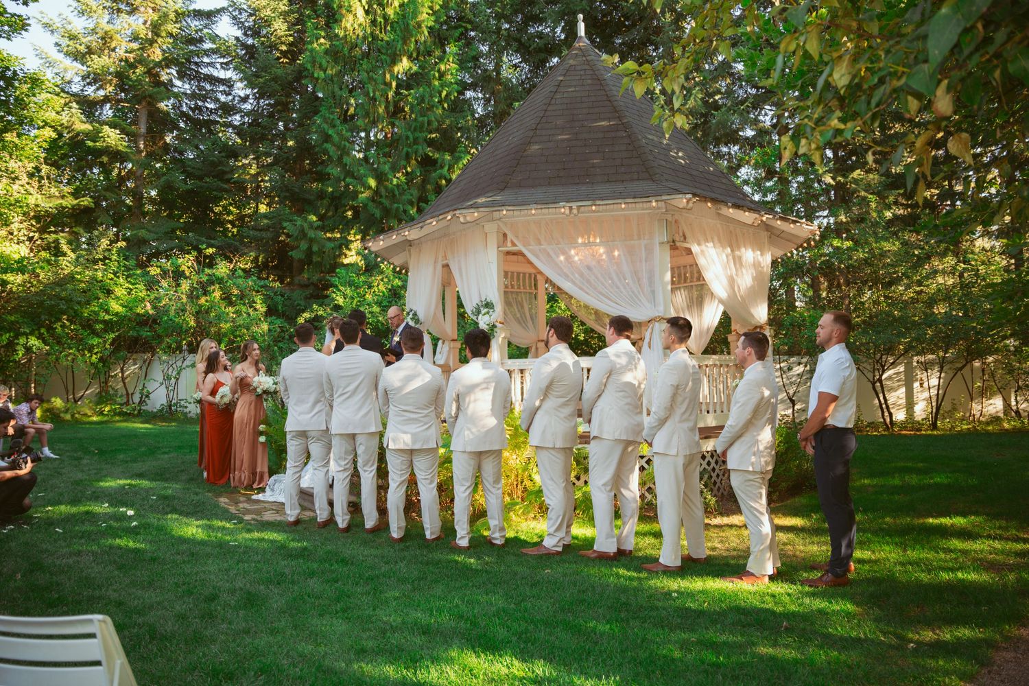 Wedding party in white attire stands beneath an elegant outdoor gazebo decorated with flowing white fabric.