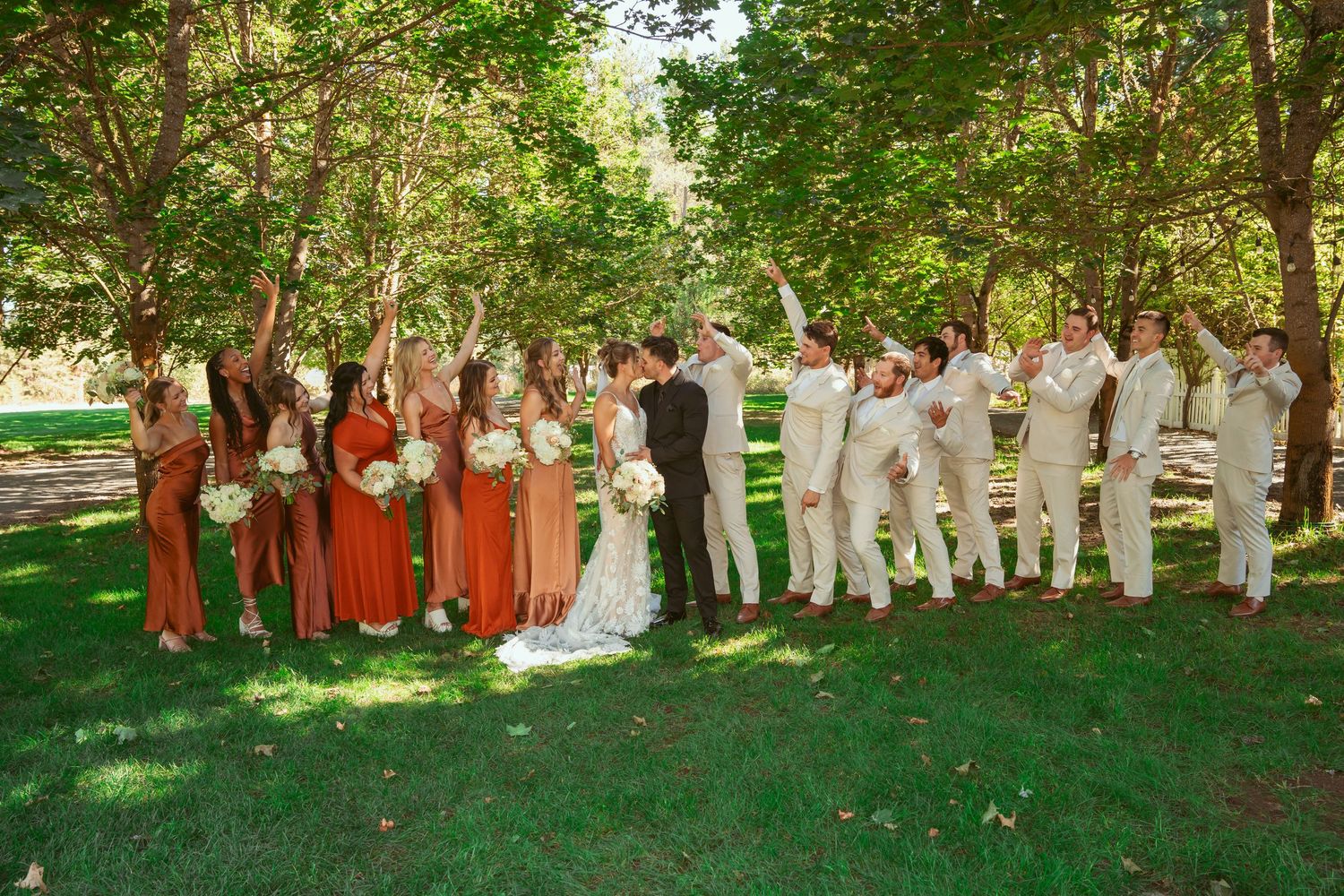 Wedding party lined up outdoors wearing orange dresses and white attire against a natural backdrop.