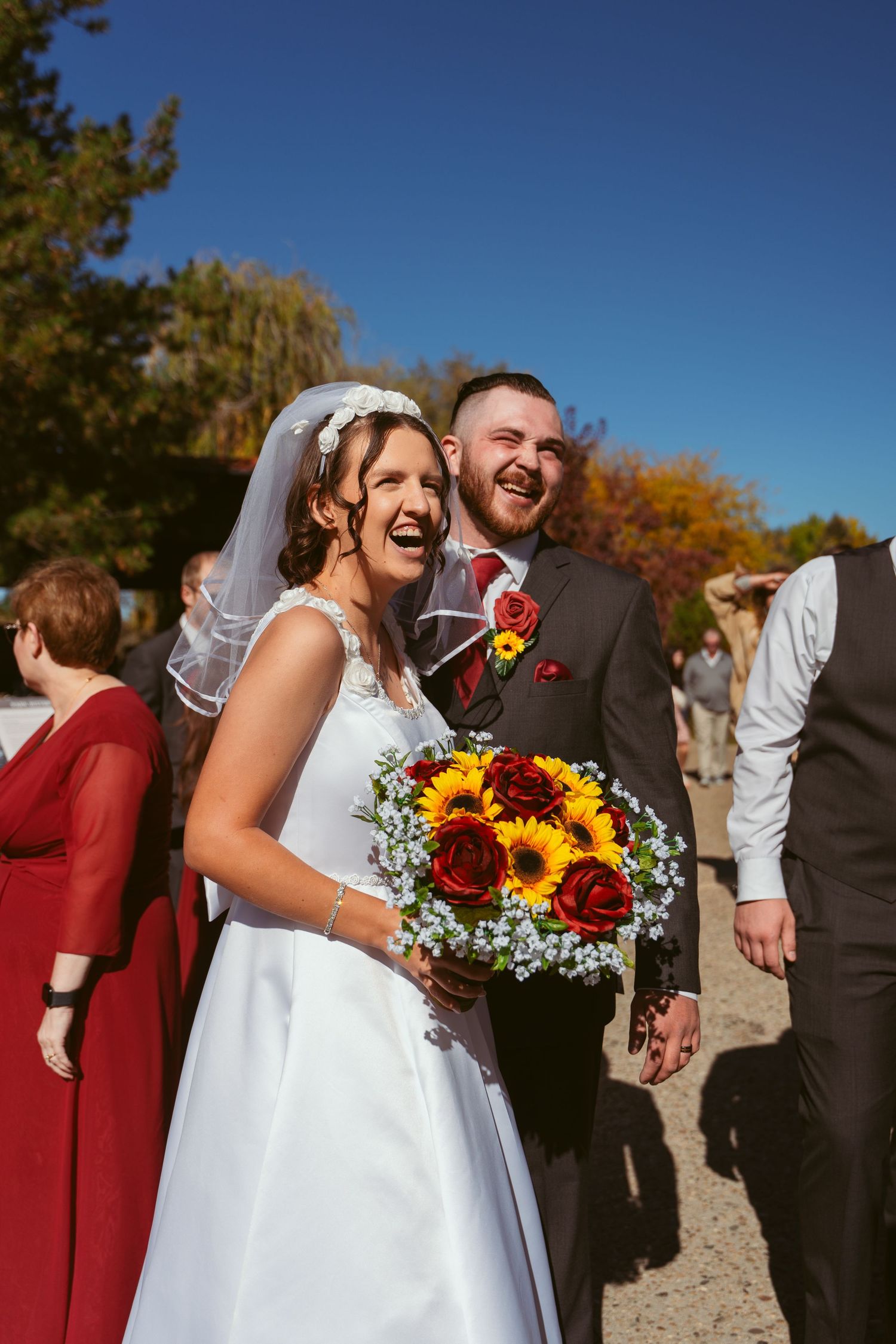 Wedding couple shares joyful moment outdoors with sunflowers and red bridesmaid dresses in background.
