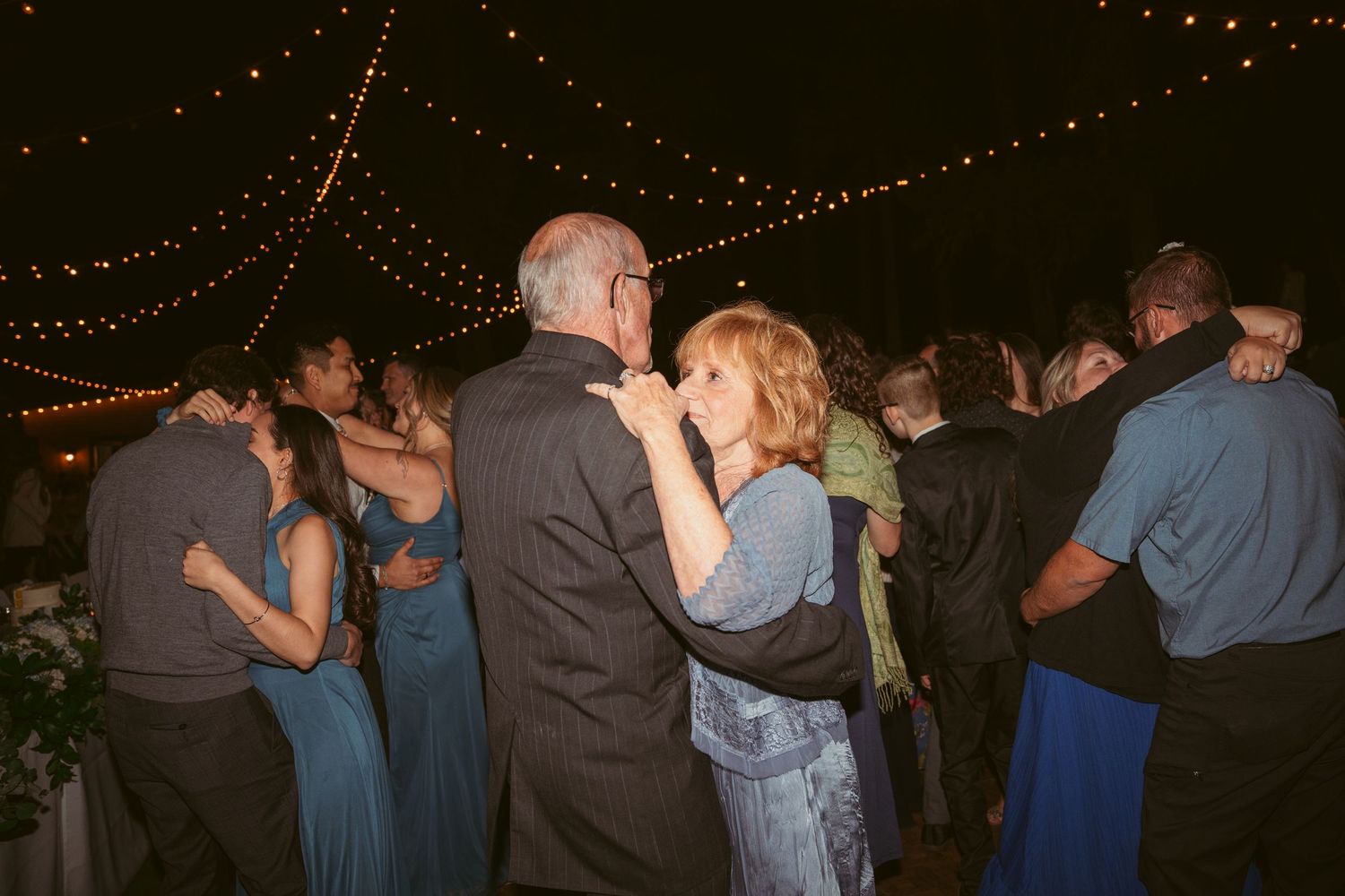 Couples slow dancing under romantic string lights at an evening outdoor event.