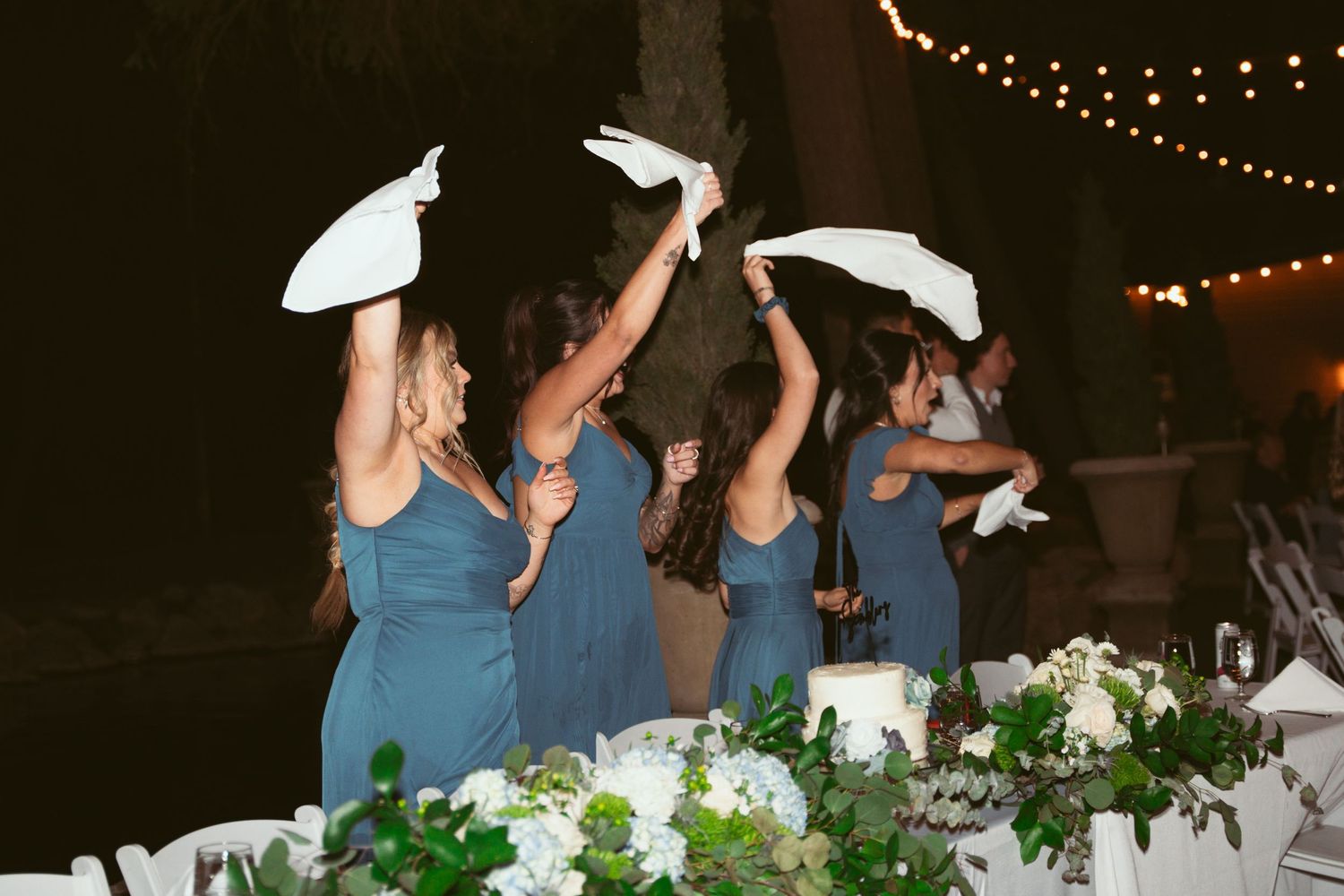 Bridesmaids in blue dresses dance together at an outdoor wedding reception.