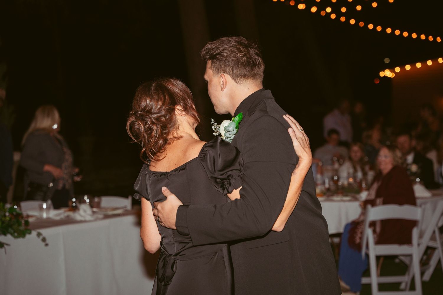 Wedding guests dance and celebrate on a dark outdoor dance floor with string lights above.
